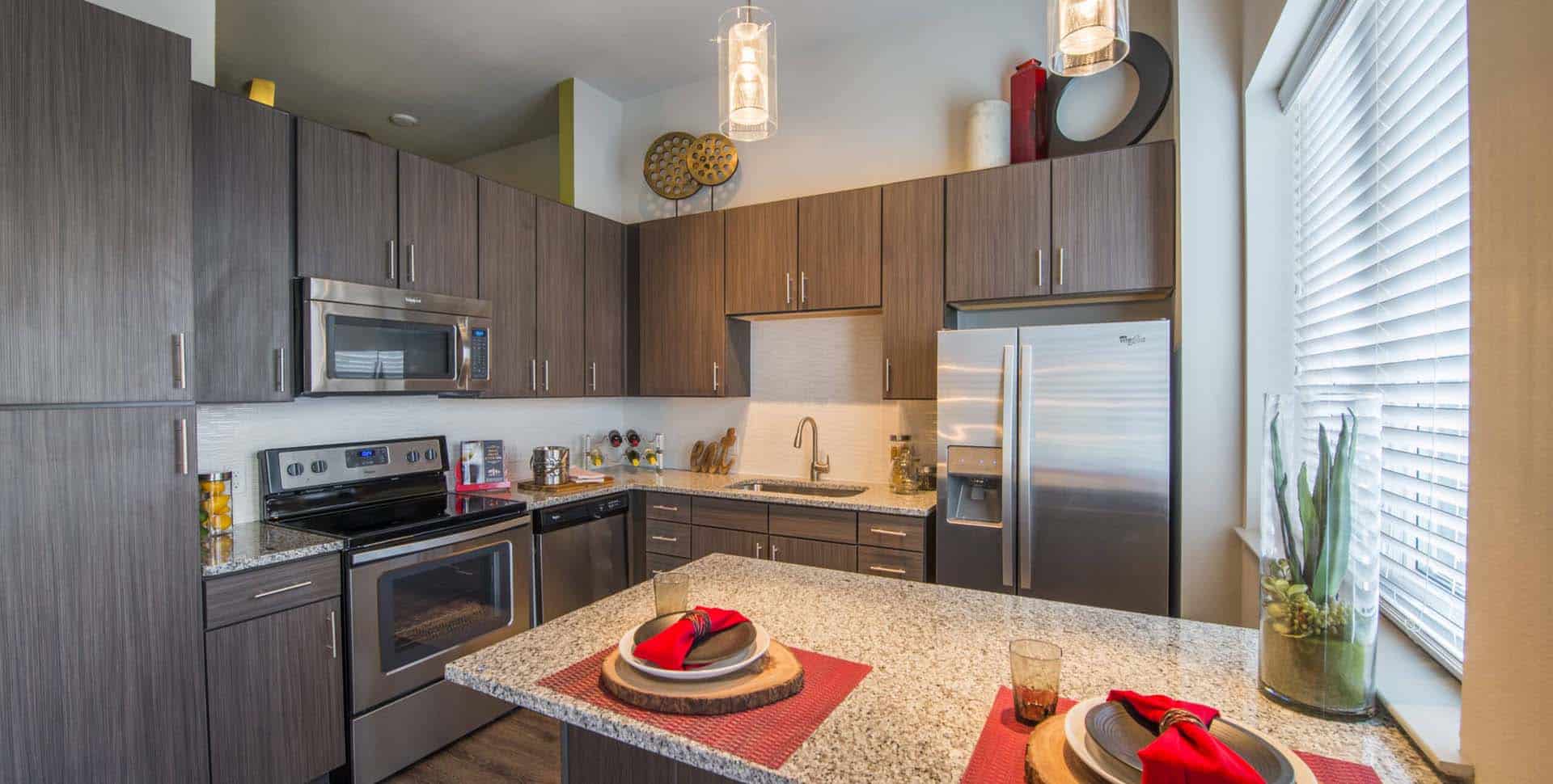 Modern kitchen with stainless steel appliances, brown cabinets, granite countertops, and place settings for two on the island; natural light from a large window.