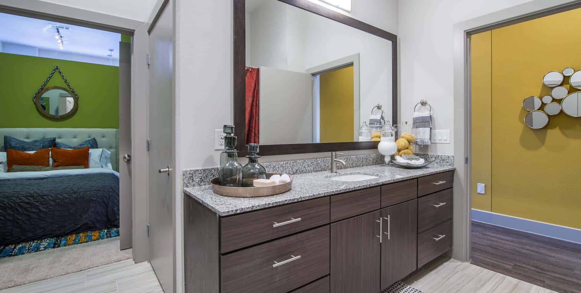 Modern bathroom with a large mirror, granite countertop, and dark wood cabinets, adjacent to a bedroom with a green accent wall and colorful bedding.