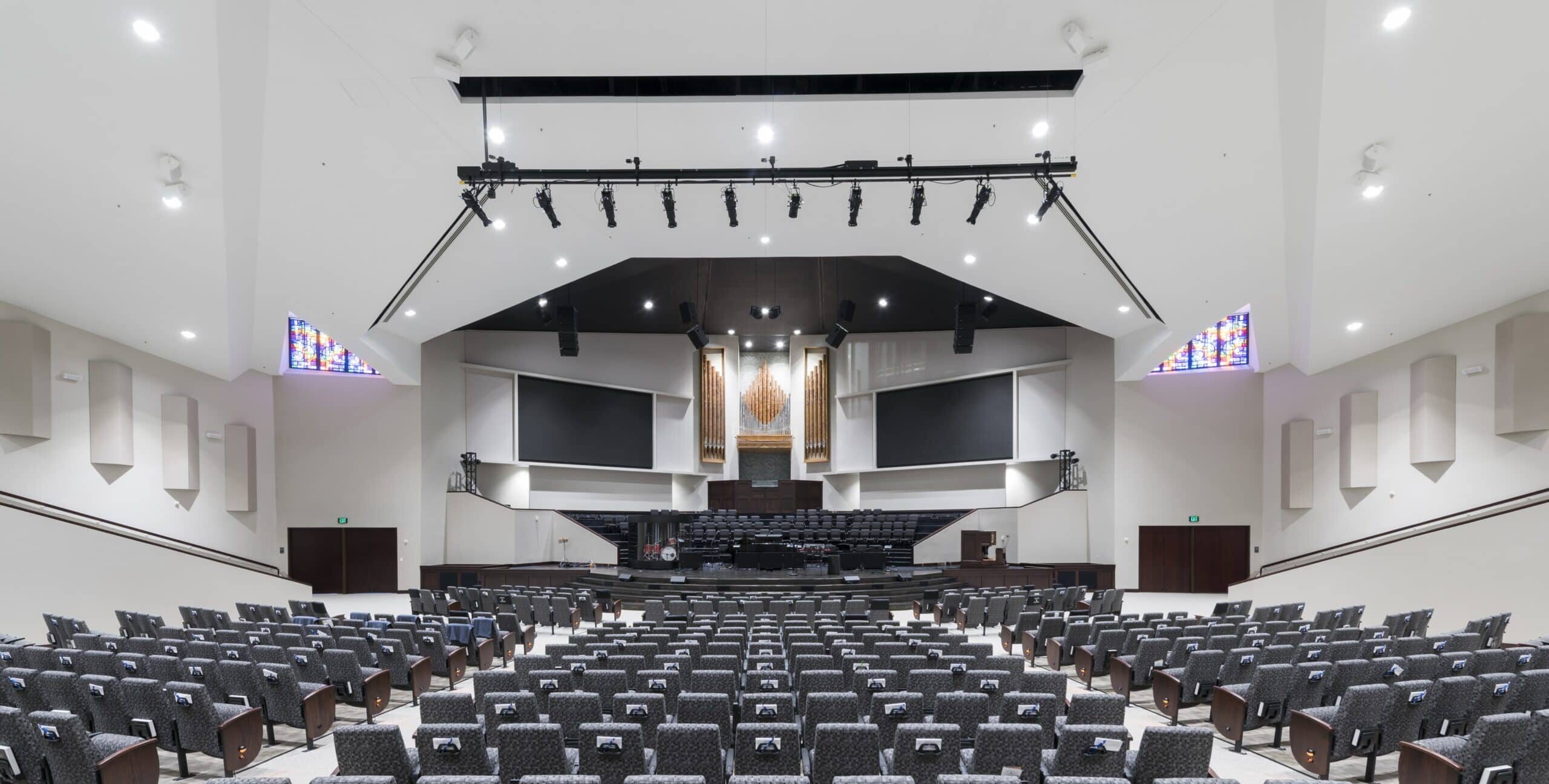 A large auditorium or sanctuary with semicircular rows of cushioned seats facing a stage equipped with multiple screens, musical instruments, and wooden decorative panels. Overhead lighting and stained‑glass windows line the upper walls.