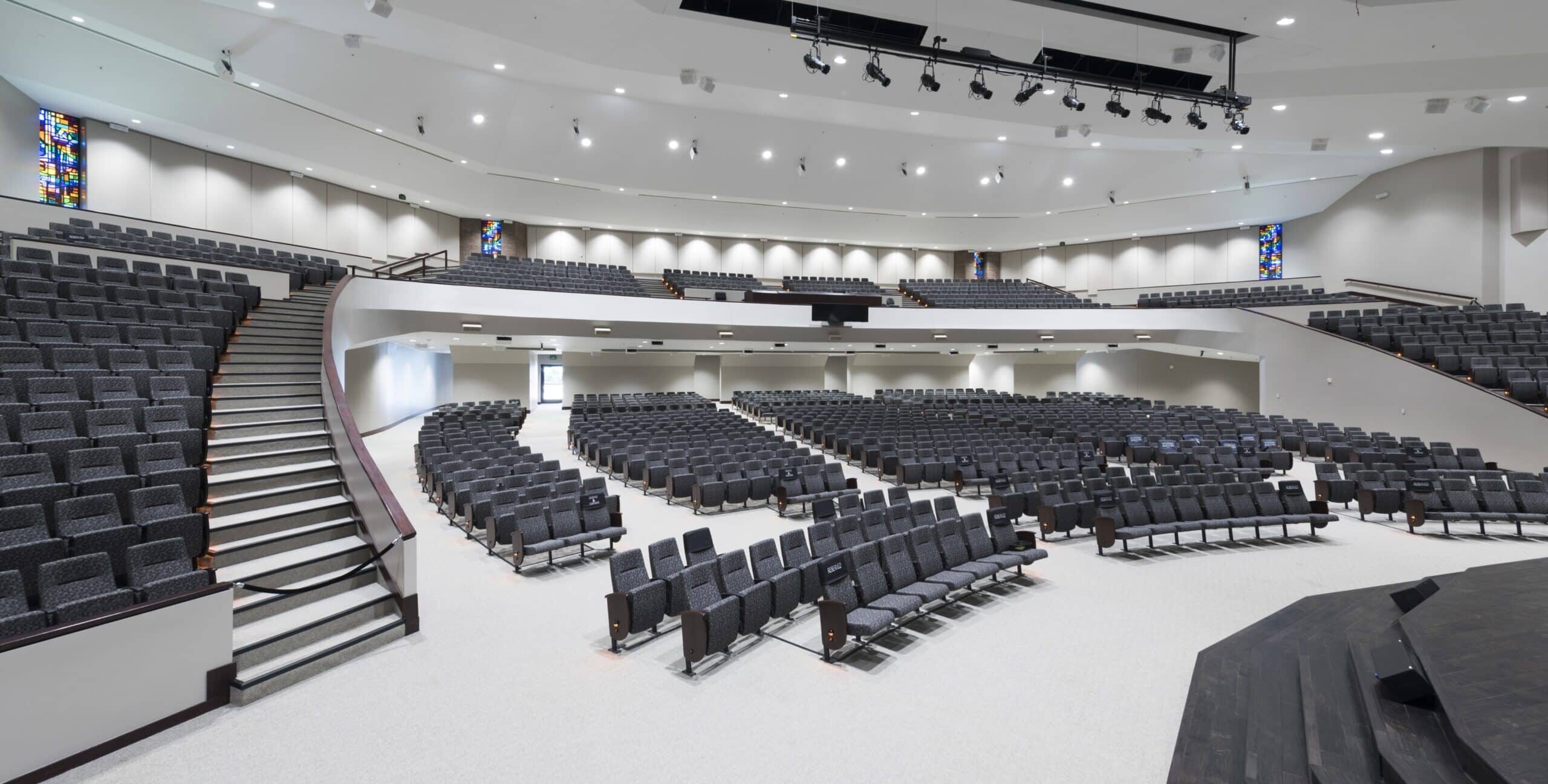 A large modern auditorium with tiered semicircular seating facing a stage. Dark gray upholstered chairs, stained‑glass windows along the side walls, and overhead stage lighting create a clean, contemporary performance space.