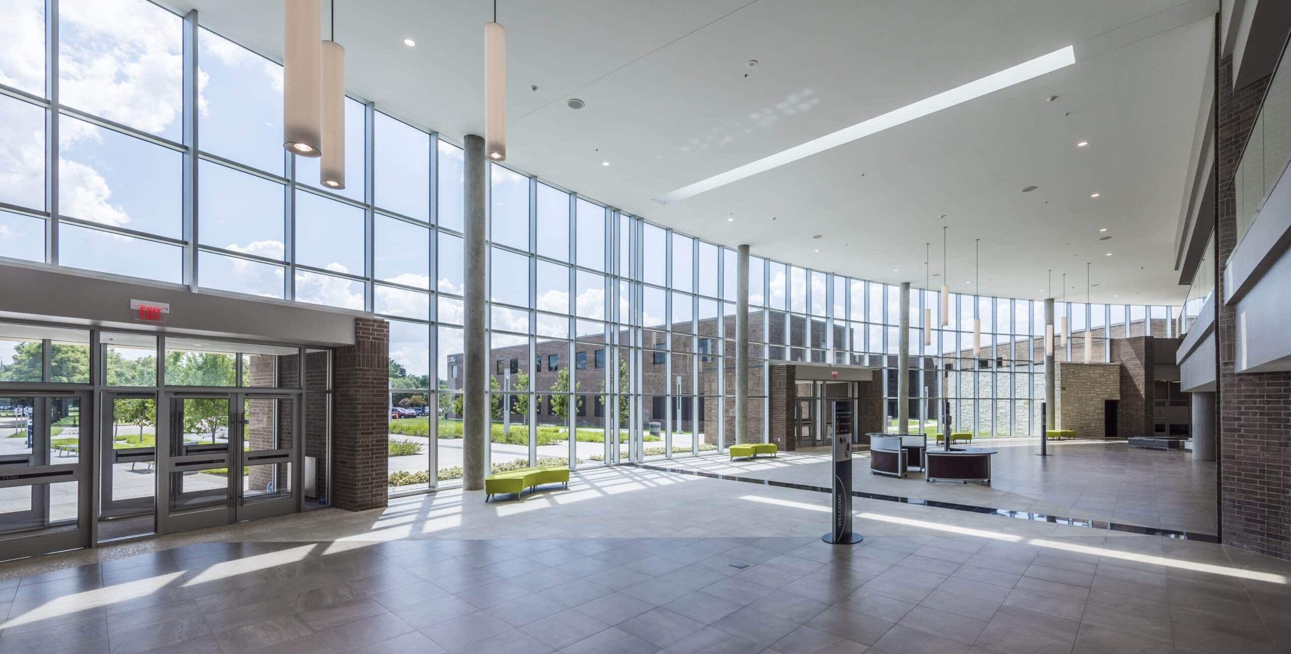 A bright lobby with tall glass windows, gray tile flooring, and cylindrical hanging lights. A circular reception desk sits near the center, with green benches along the windows and landscaped greenery visible outside.