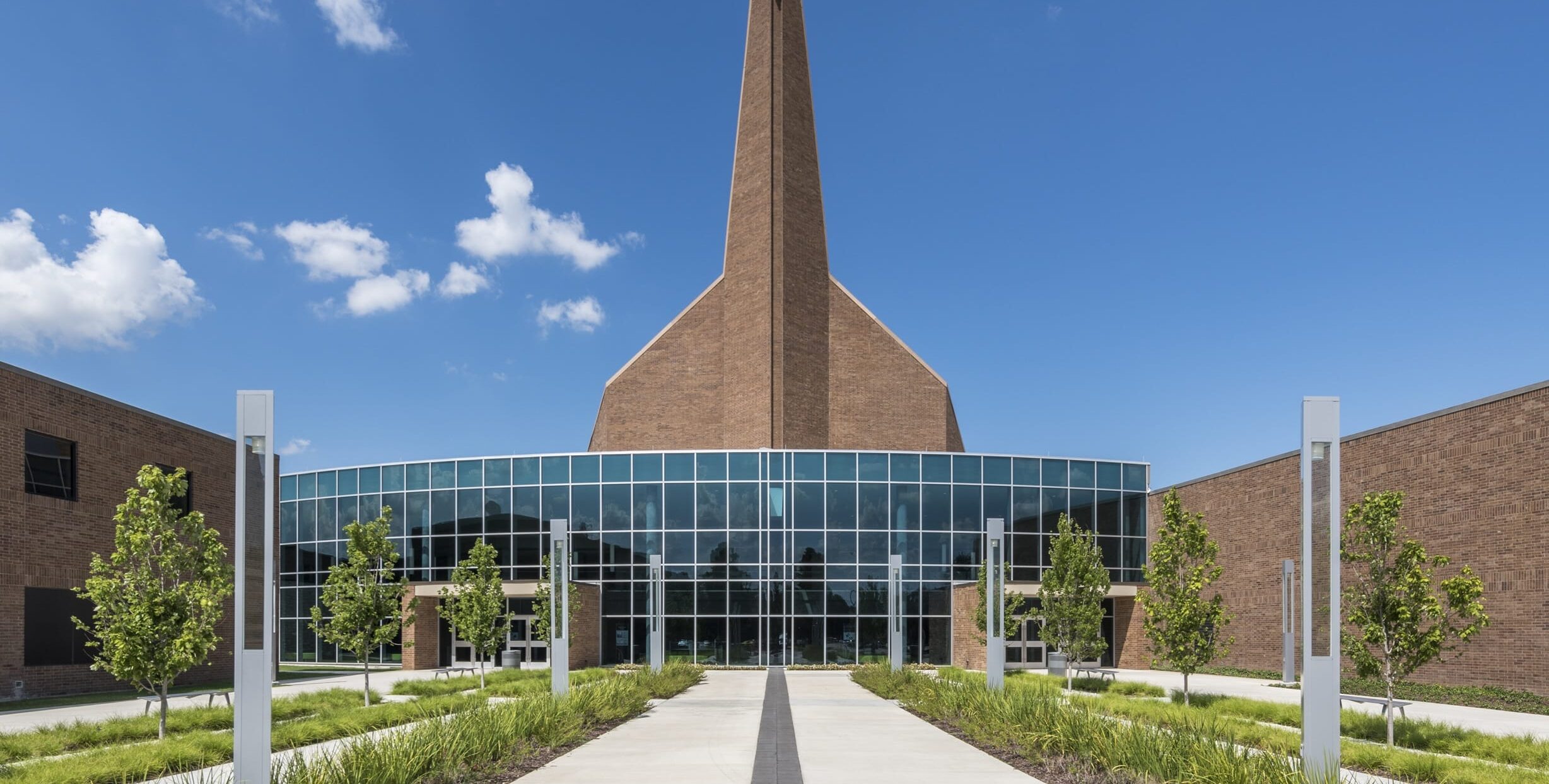 A modern church exterior with a tall brick steeple topped by a white cross. Large grid‑patterned windows reflect the sky above a wide concrete walkway lined with grass, small trees, and decorative light posts.