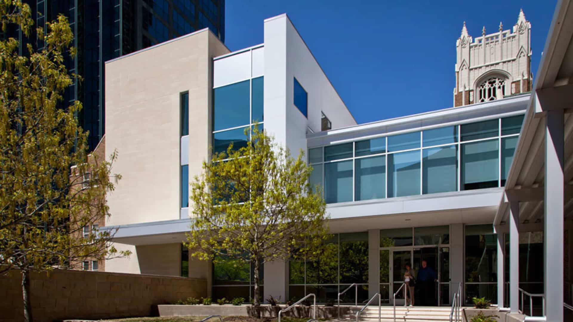 Modern building with large glass windows and a stone facade, surrounded by trees, with a historic tower visible in the background under a clear blue sky.