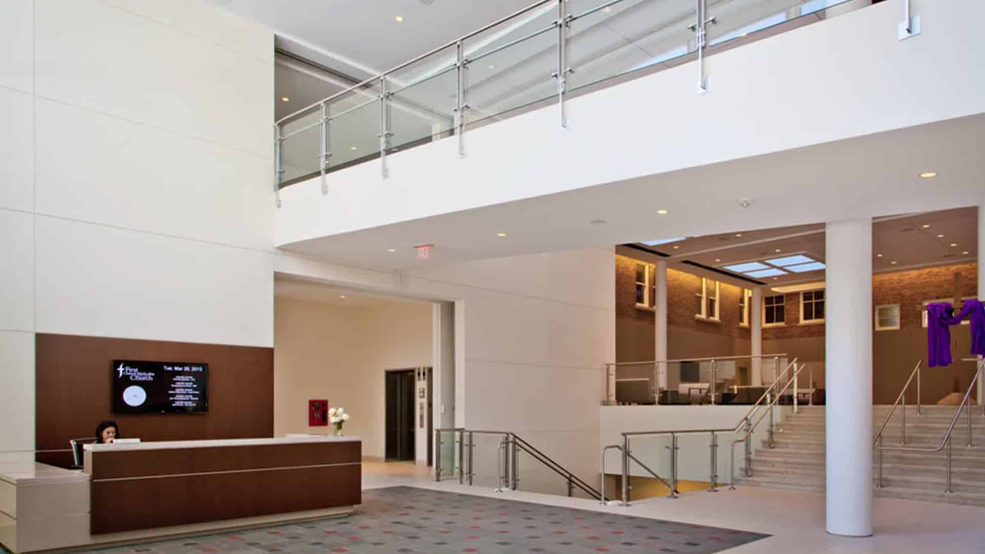 Modern lobby area with a reception desk, staircase, glass railings, high ceilings, and natural light from large windows.