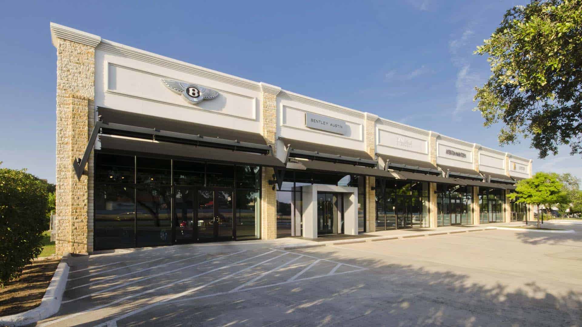 A modern commercial building with large glass windows and several retail storefronts, including a Bentley dealership, on a sunny day.