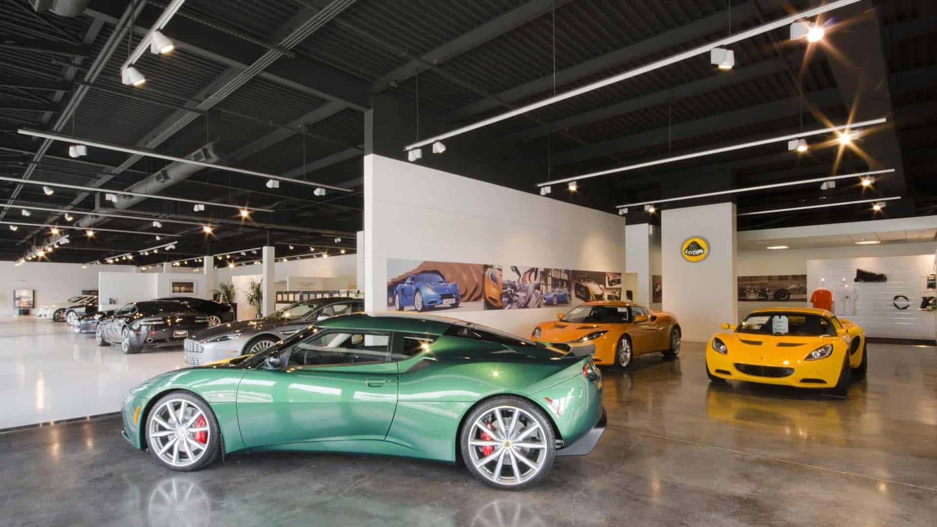 A modern car showroom displays several sports cars, including a green, an orange, and a yellow car, under bright ceiling lights.