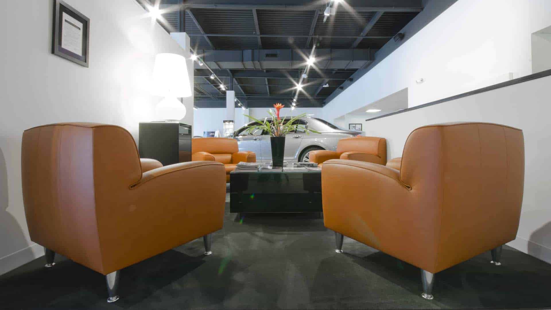Four orange armchairs surround a black coffee table with a flower vase in a modern waiting area; a silver car is visible in the background.