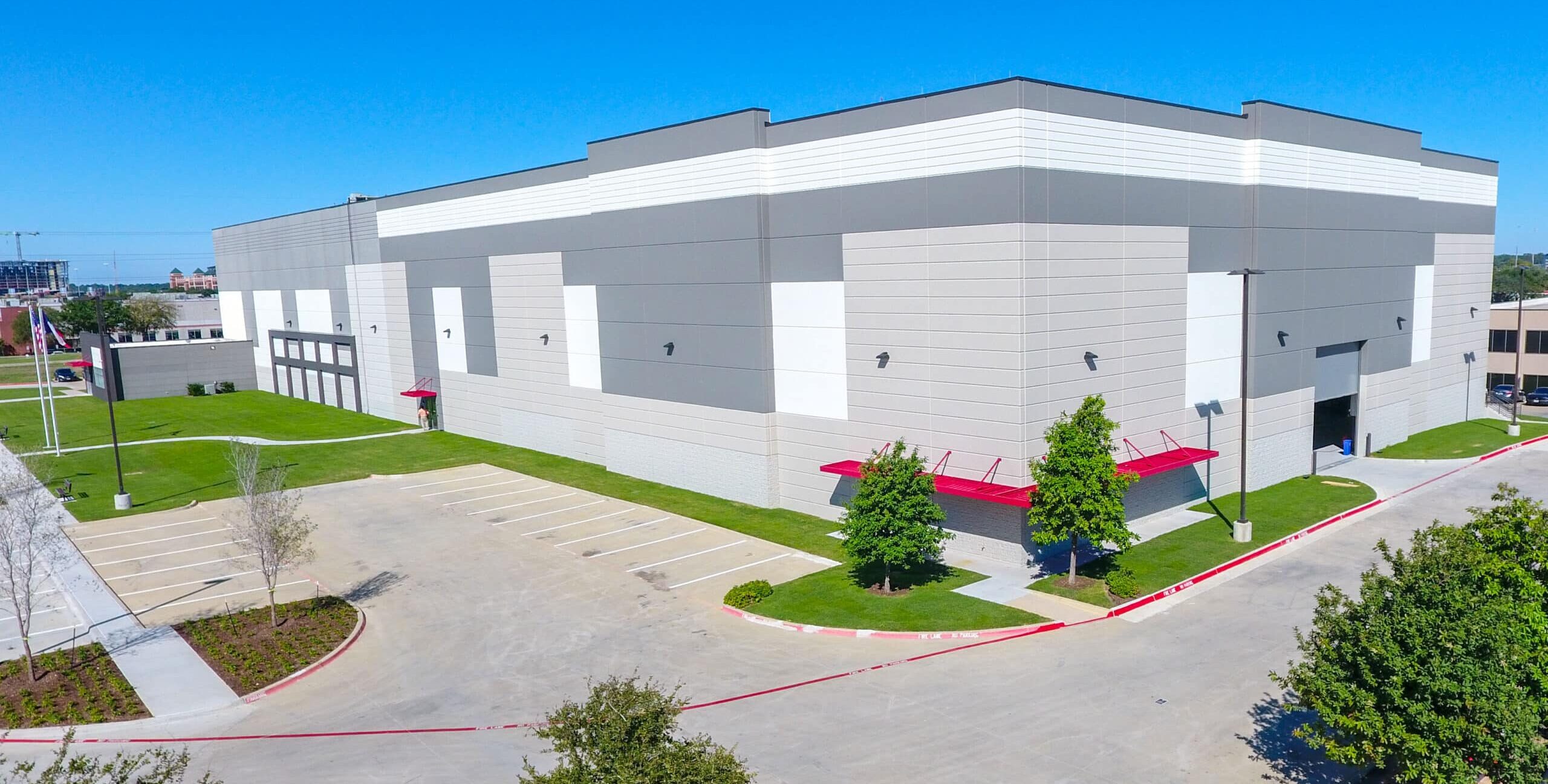 Large, modern industrial warehouse with gray and white exterior, red awnings, empty parking lot, and a few trees under a clear blue sky.