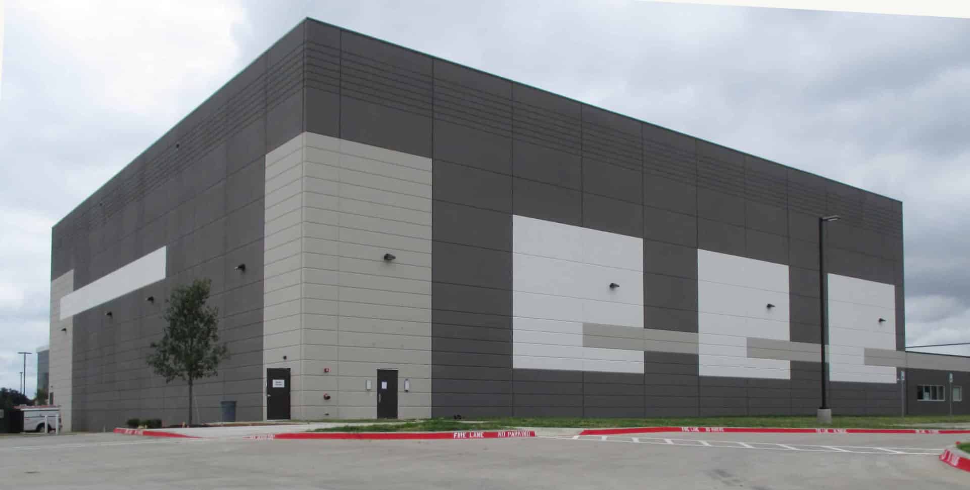 A large, windowless industrial building with geometric patterns on the exterior walls, surrounded by a paved parking lot and a few small trees.