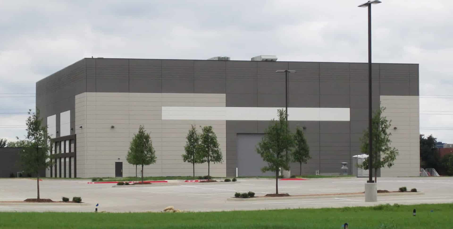 A large, rectangular industrial building with gray and white exterior panels, surrounded by a mostly empty parking lot and a few small trees.