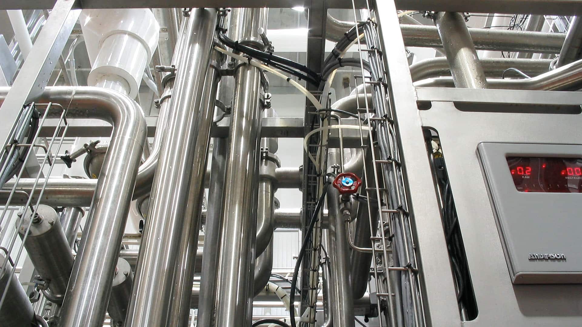 Close-up view of industrial stainless steel pipes, valves, and a control panel with digital readouts in a factory or processing facility.
