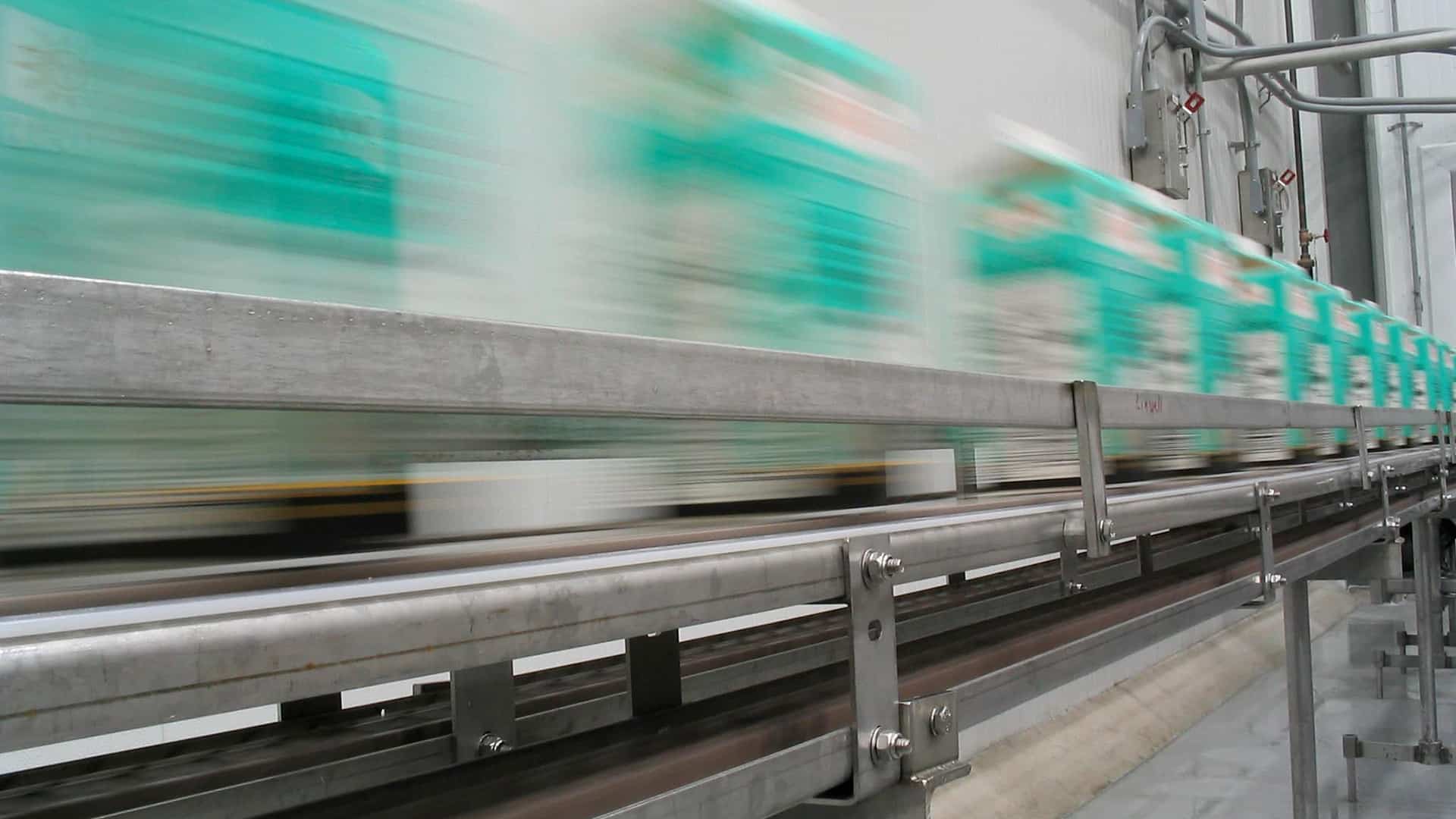Green and white cartons move rapidly along a conveyor belt in a factory setting, creating a motion blur effect.