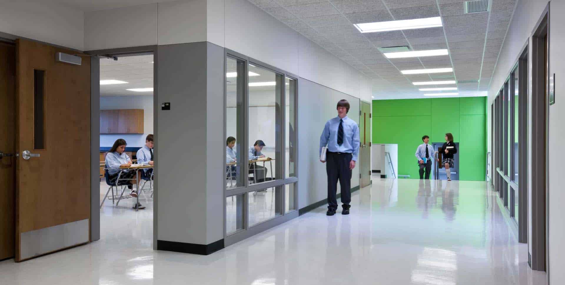 Students in uniforms sit in a classroom while others walk in a bright, modern school hallway with white floors and green accent walls.