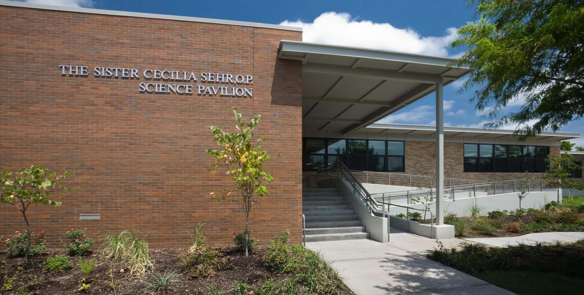 A brick building labeled "The Sister Cecilia Sehr, O.P. Science Pavilion" has a ramp and stairs leading to the entrance, with landscaped plants in front.