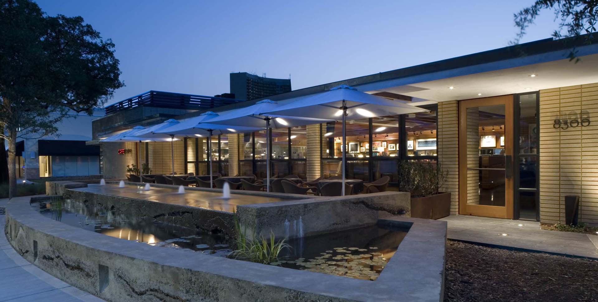 Modern restaurant exterior at dusk with large windows, patio seating under umbrellas, and a concrete water feature with fountains and water lilies in the foreground.