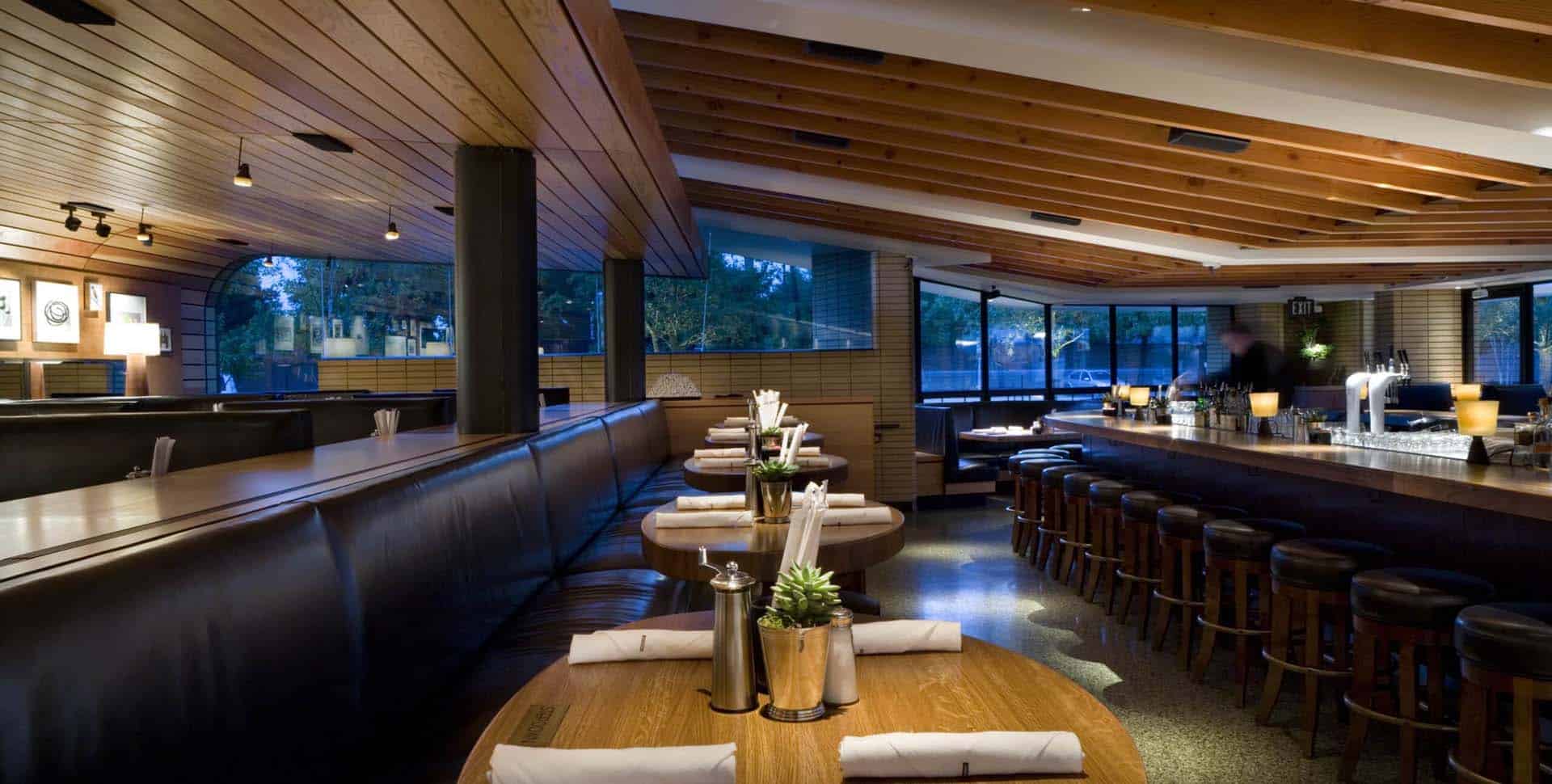 Modern restaurant interior with wooden ceiling, long bar lined with stools, booth seating, and tables set with napkins and condiments. Large windows provide natural light.