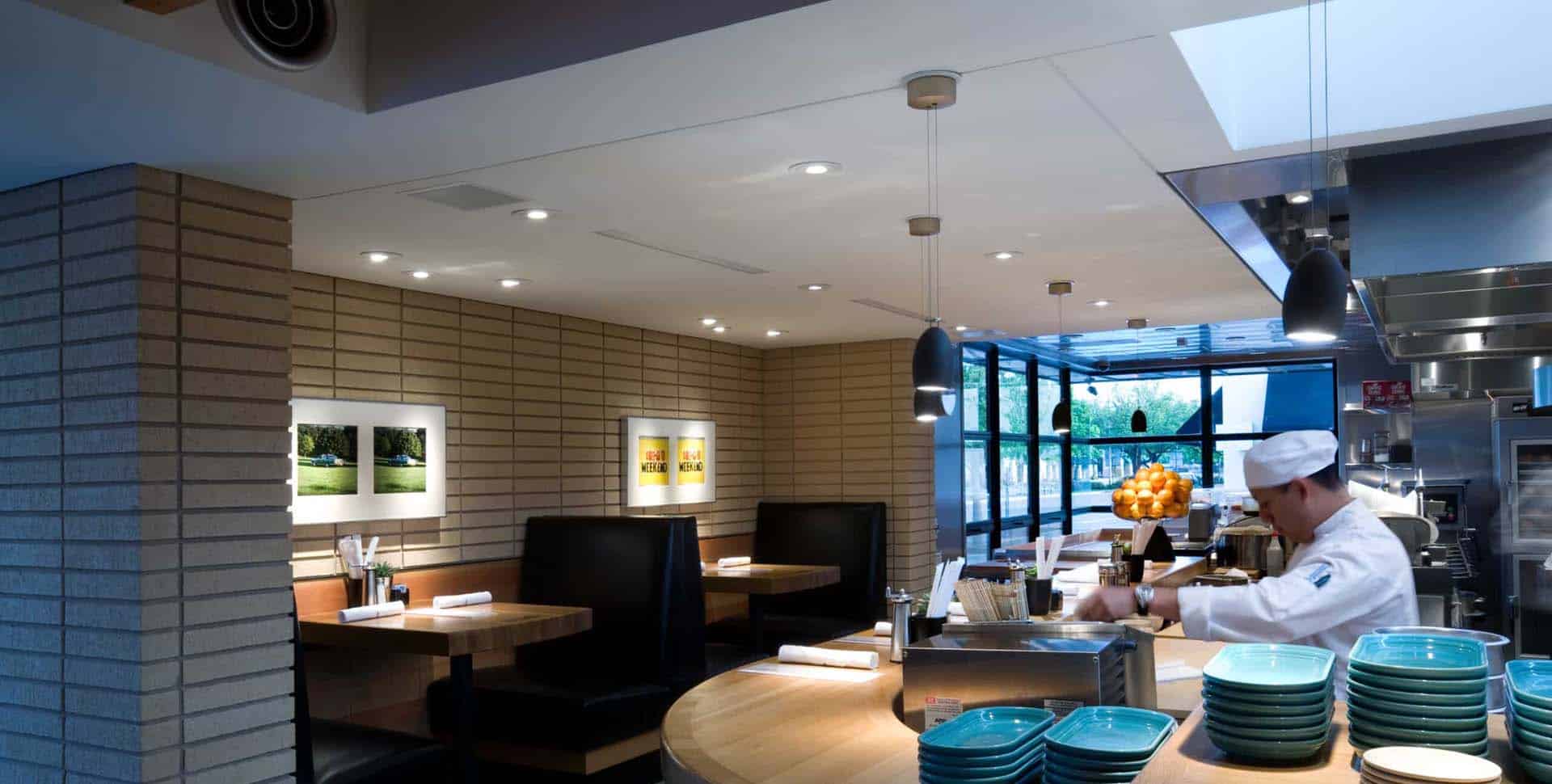 A chef prepares food behind a counter in a modern restaurant with booth seating, brick walls, artwork, and stacked blue plates.