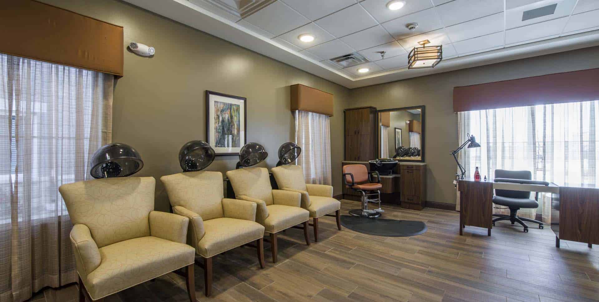 A salon room with three beige chairs and hair dryers, a styling chair at a station with a mirror, and a desk by the windows.