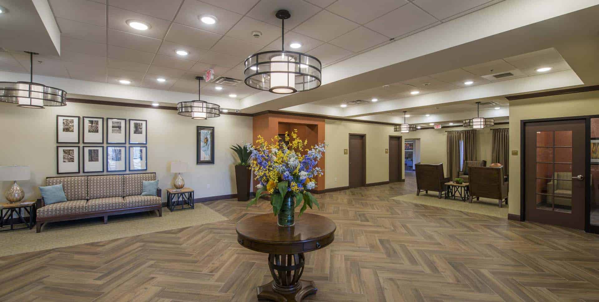 A spacious, well-lit lobby with wood-patterned floors, a round table with a flower arrangement, seating areas, and framed art on the walls.