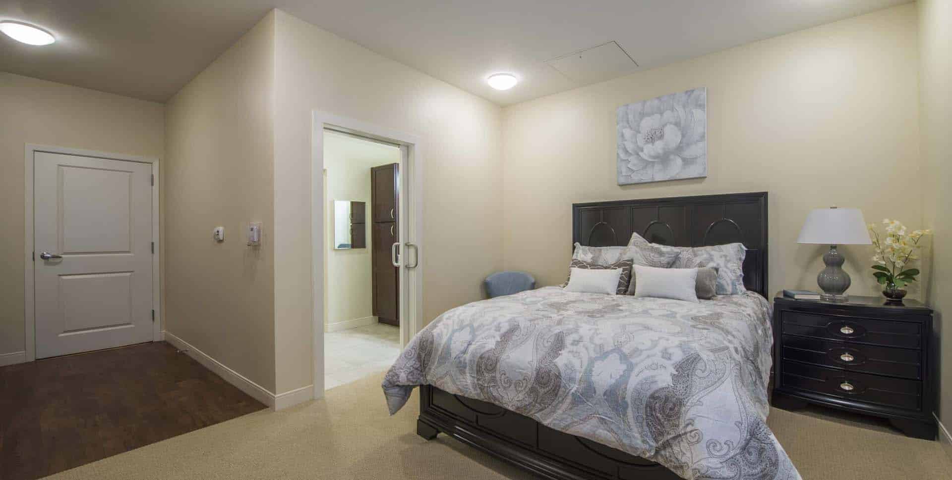 A neatly made bed with patterned bedding sits next to two nightstands in a beige bedroom, with a door leading to an adjoining bathroom.
