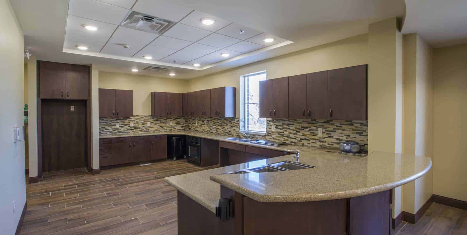 Modern kitchen with brown cabinets, tiled backsplash, and a curved island countertop. The room has recessed lighting, a window, and wood-look tile flooring.