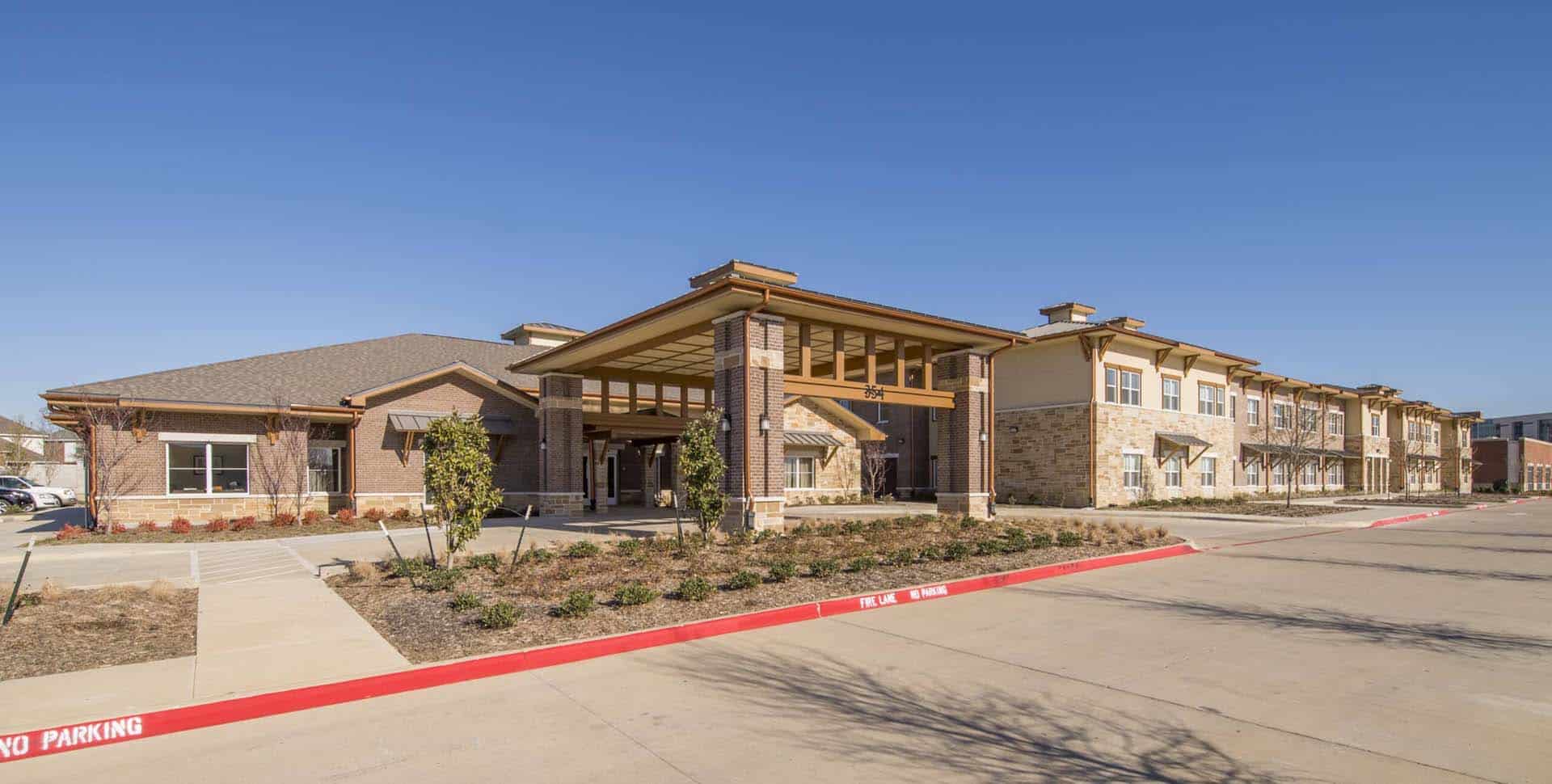 A modern two-story building with stone and brick exterior, large covered entrance, and landscaped grounds under a clear blue sky.