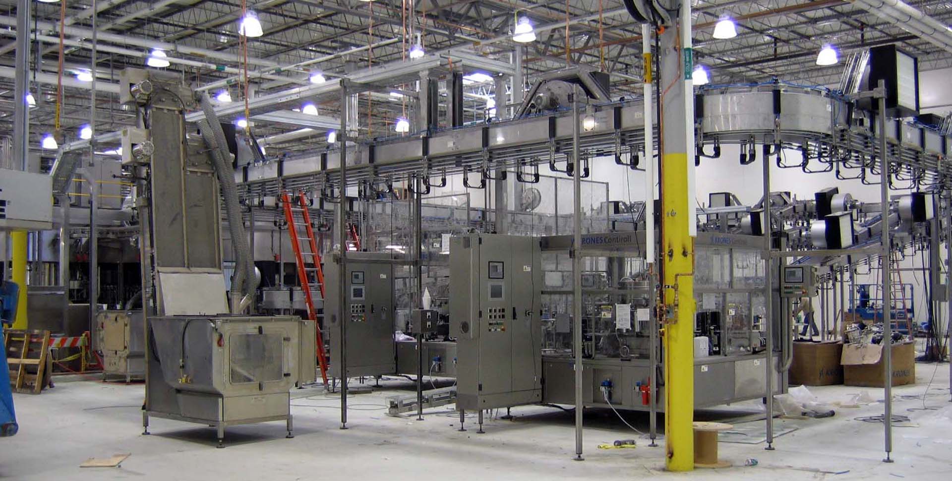 Automated bottling line in a factory with conveyor belts, metal machinery, pipes, and industrial lighting in a spacious, high-ceilinged environment.
