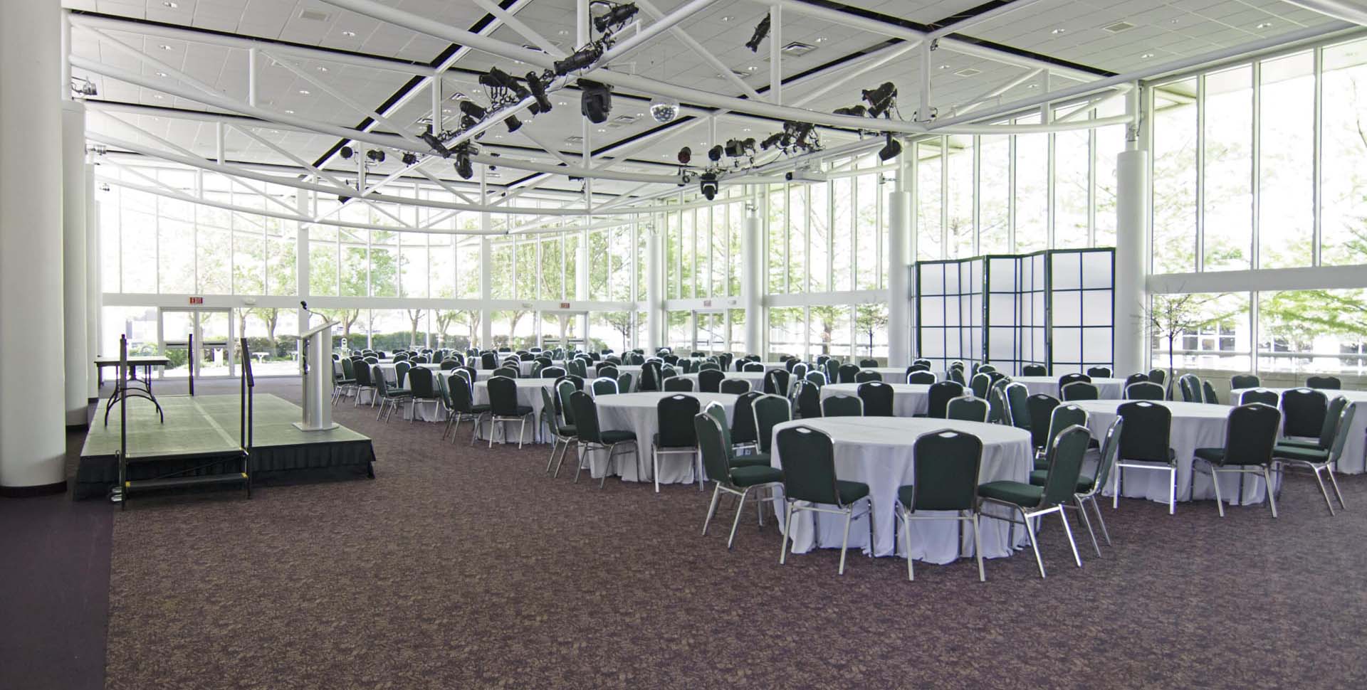 A spacious event hall with round tables and chairs arranged for a large gathering, floor-to-ceiling windows, and a podium on a small stage.