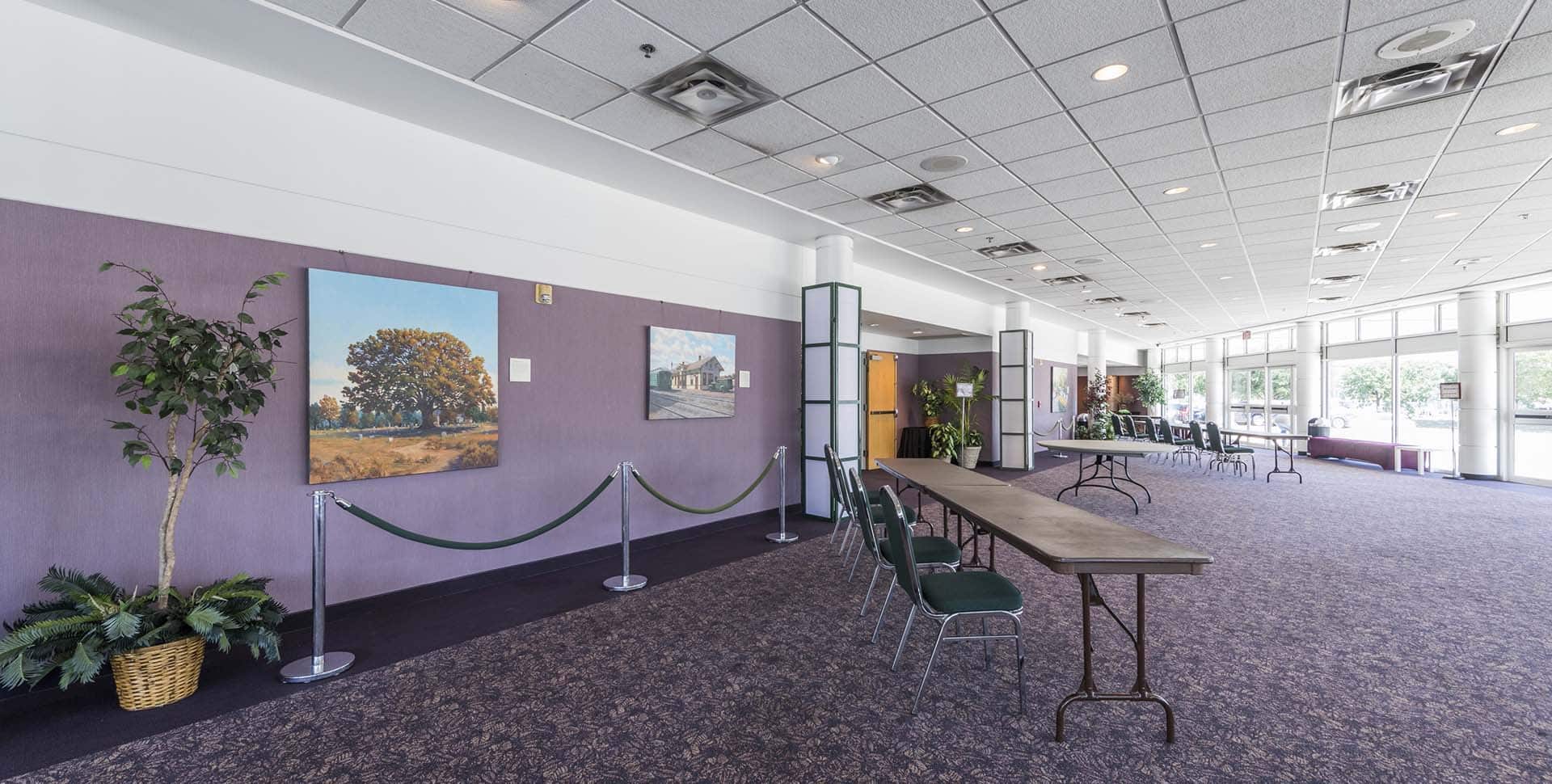 A spacious lobby with purple walls, a long table with green chairs, rope barriers, potted plant, and landscape paintings on the wall. Large windows let in natural light.