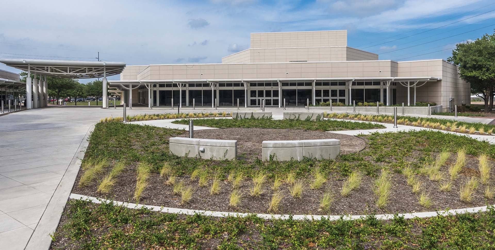 Modern, beige building with large windows and landscaped circular garden in front, featuring concrete pathways and ornamental grasses under a partly cloudy sky.