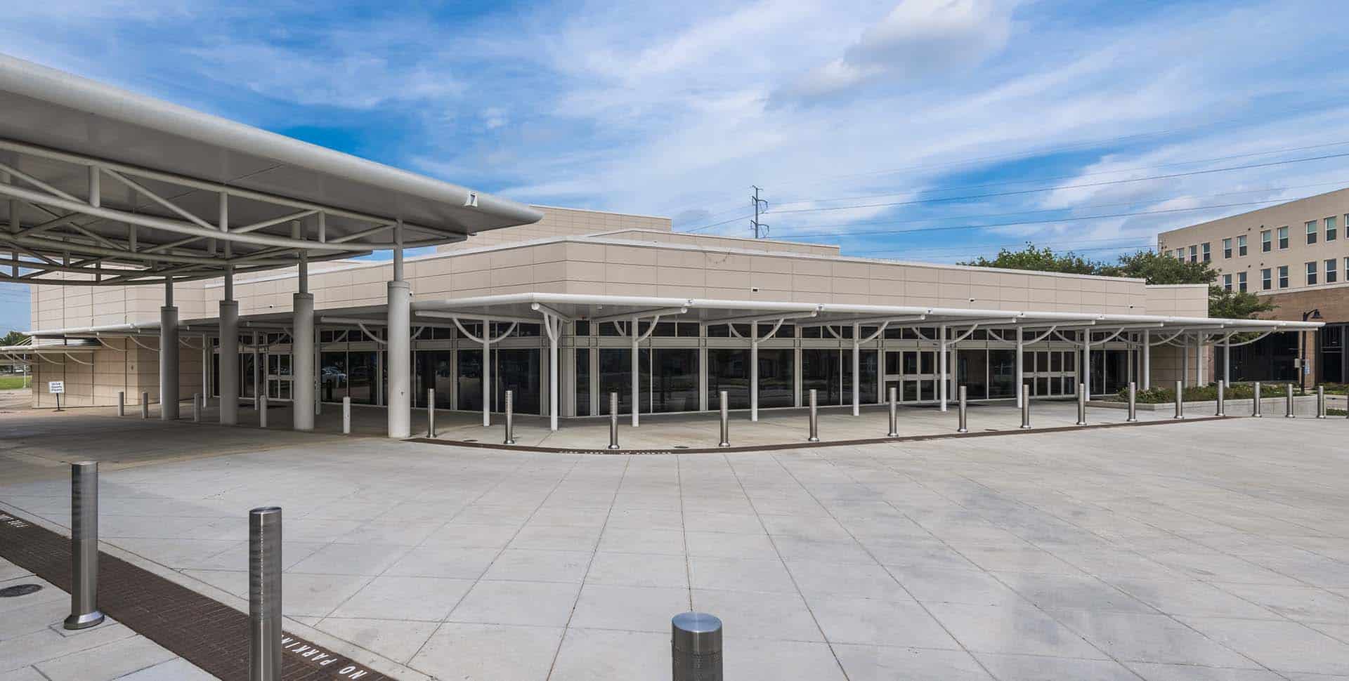A modern building with large windows, multiple entrances, and a covered walkway on a wide, paved area under a partly cloudy sky.