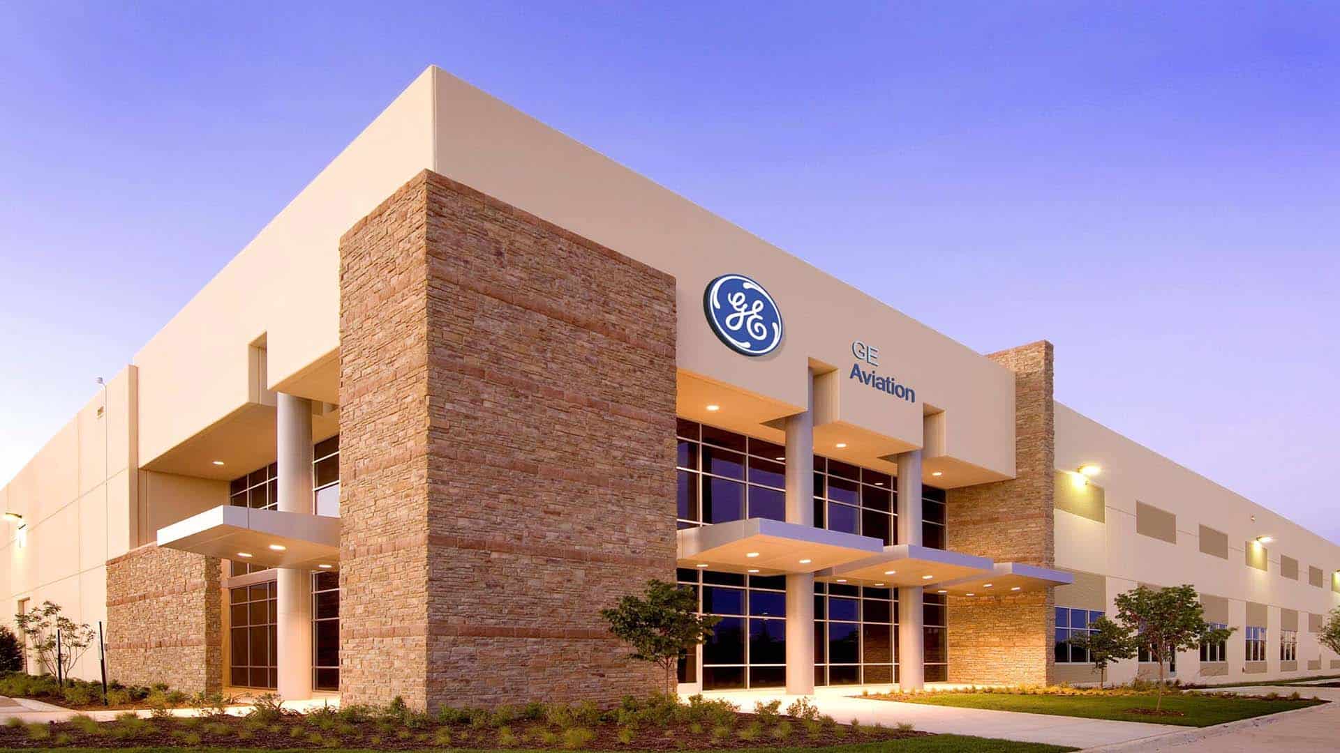Modern building with a stone and glass facade featuring the GE Aviation logo and sign, surrounded by landscaped shrubs and trees under a clear evening sky.