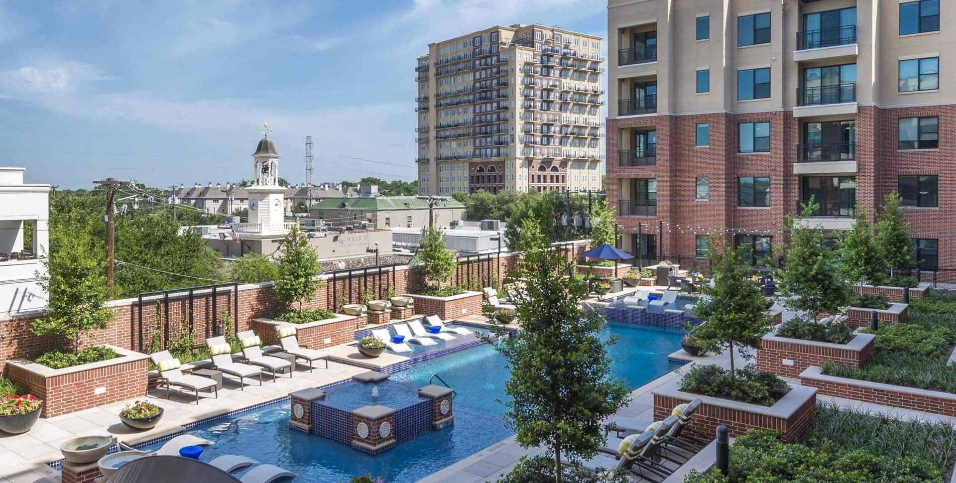 Modern apartment complex with a swimming pool, lounge chairs, landscaped greenery, and nearby mid-rise buildings under a clear blue sky.