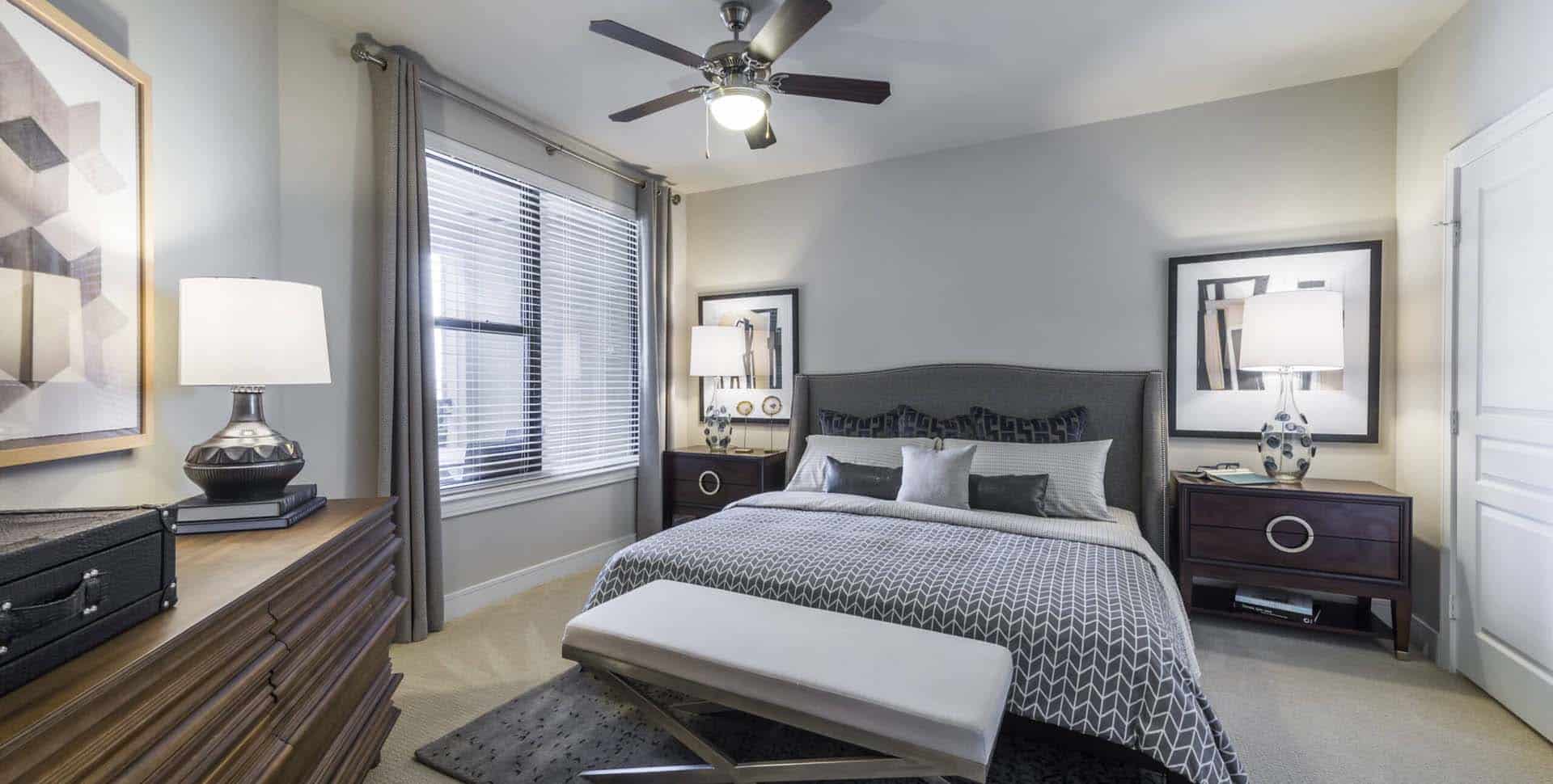 Modern bedroom with a neatly made bed, two bedside tables with lamps, large window with blinds, ceiling fan, and abstract wall art.