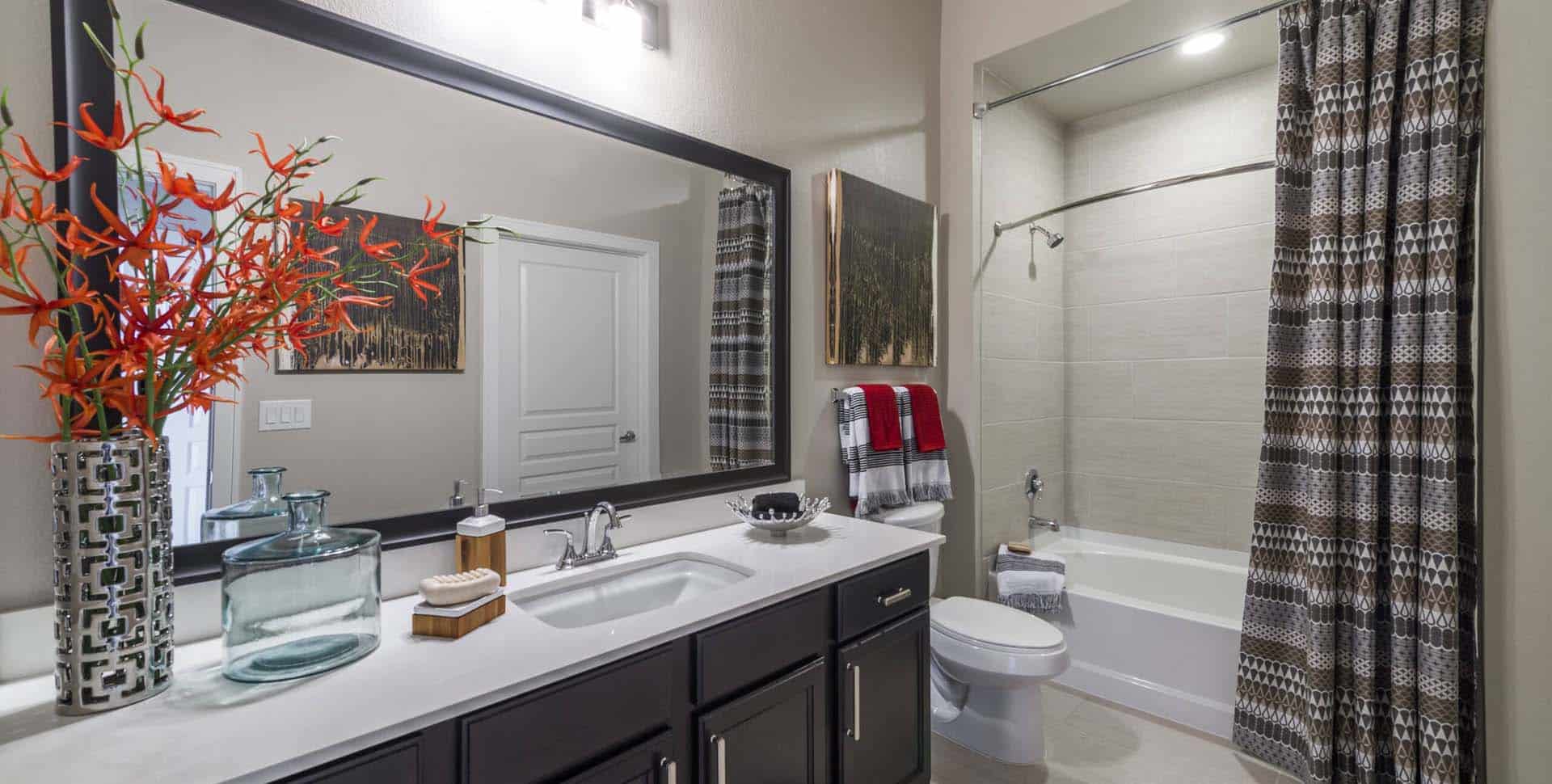 Modern bathroom with a double-sink vanity, large mirror, decorative vases, a bathtub with a patterned shower curtain, and folded towels on a rack.