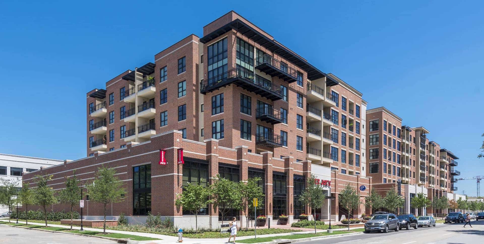 A modern mid-rise apartment building with brick exterior, balconies, and ground-floor retail, surrounded by trees and sidewalks on a clear day.