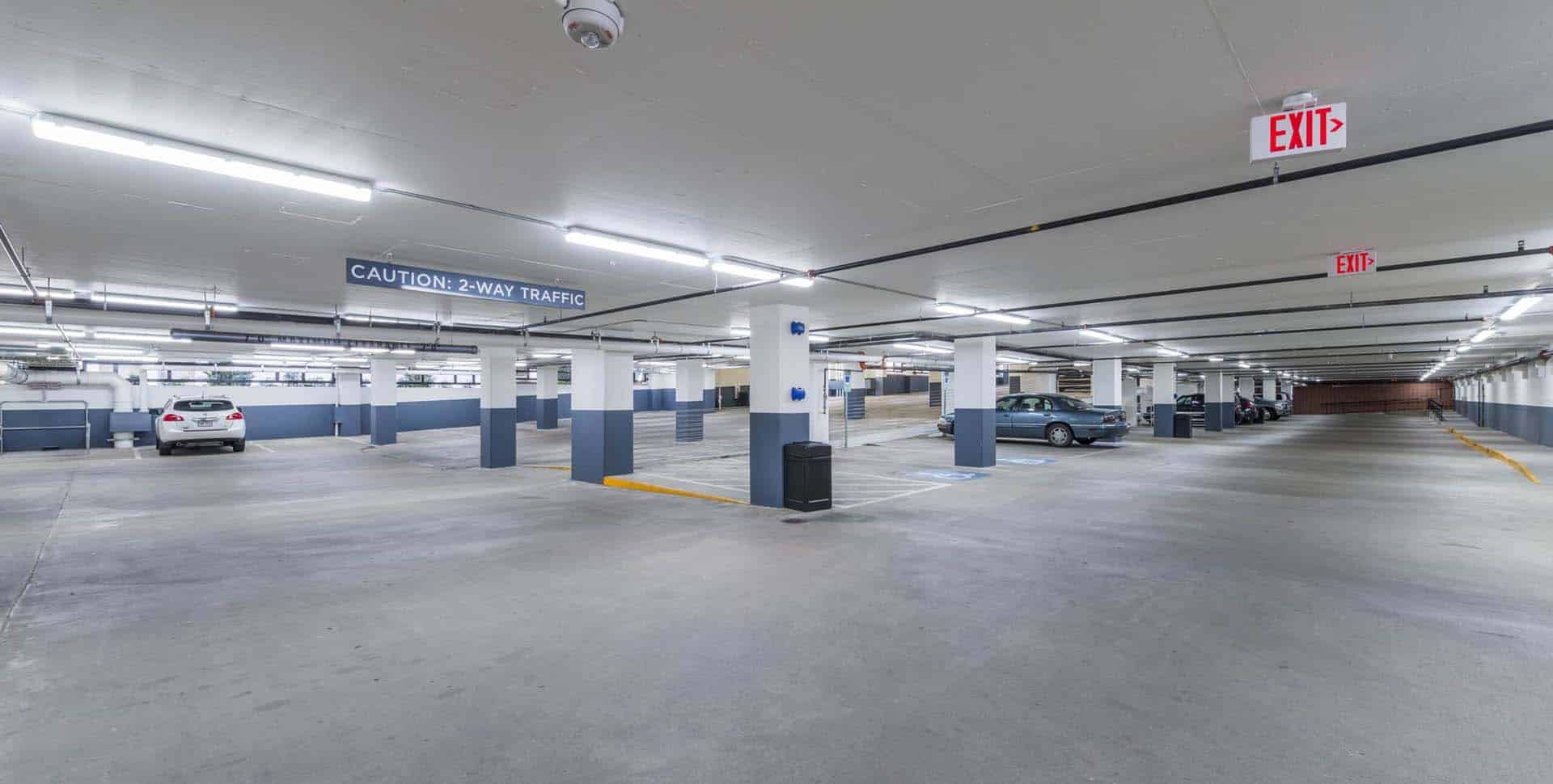 Underground parking garage with parked cars, white pillars, overhead lighting, and exit signs visible. A sign reads "Caution: 2-Way Traffic.