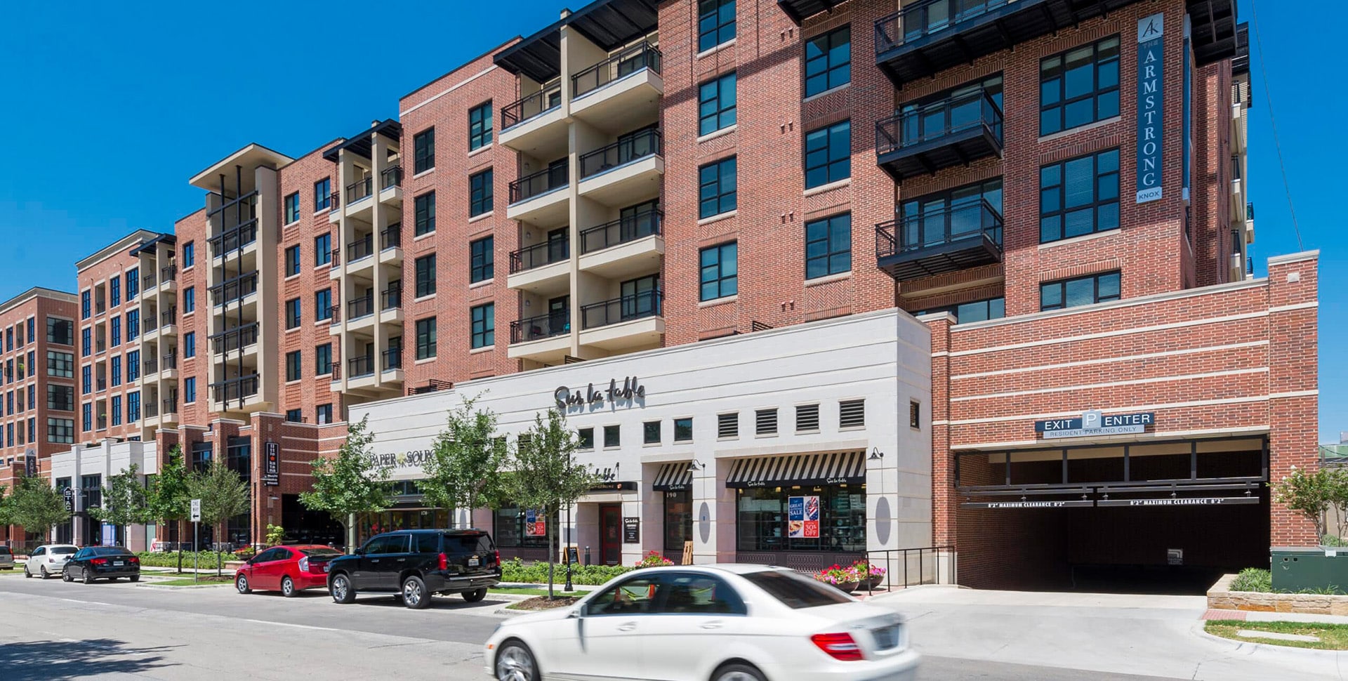 A multi-story brick apartment building with balconies and ground floor retail shops, cars parked and driving along the street in front.