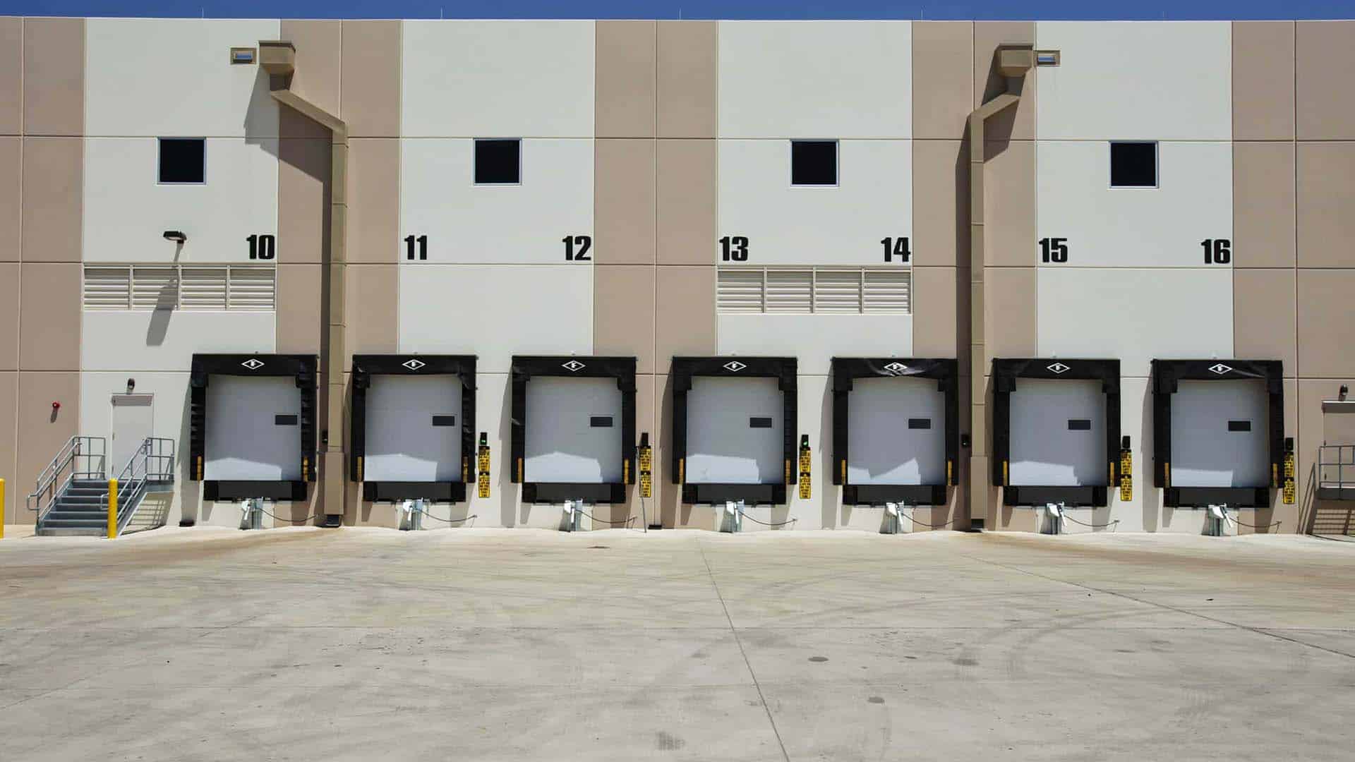 Five loading dock doors numbered 11 to 15 on a beige industrial warehouse building, with closed bay doors and an empty concrete lot in front.