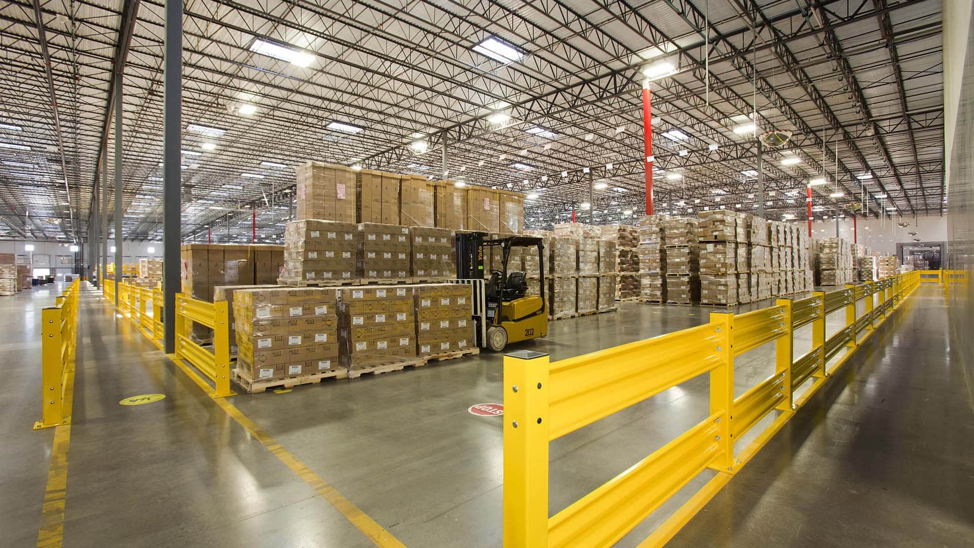 A large warehouse with stacked pallets and boxes, a forklift in operation, yellow safety barriers, and high industrial ceilings with overhead lighting.