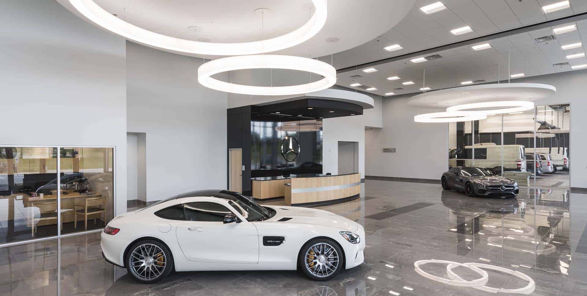 A modern car showroom with a white sports car and a black sedan on display, large circular ceiling lights, and a reception desk in the background.