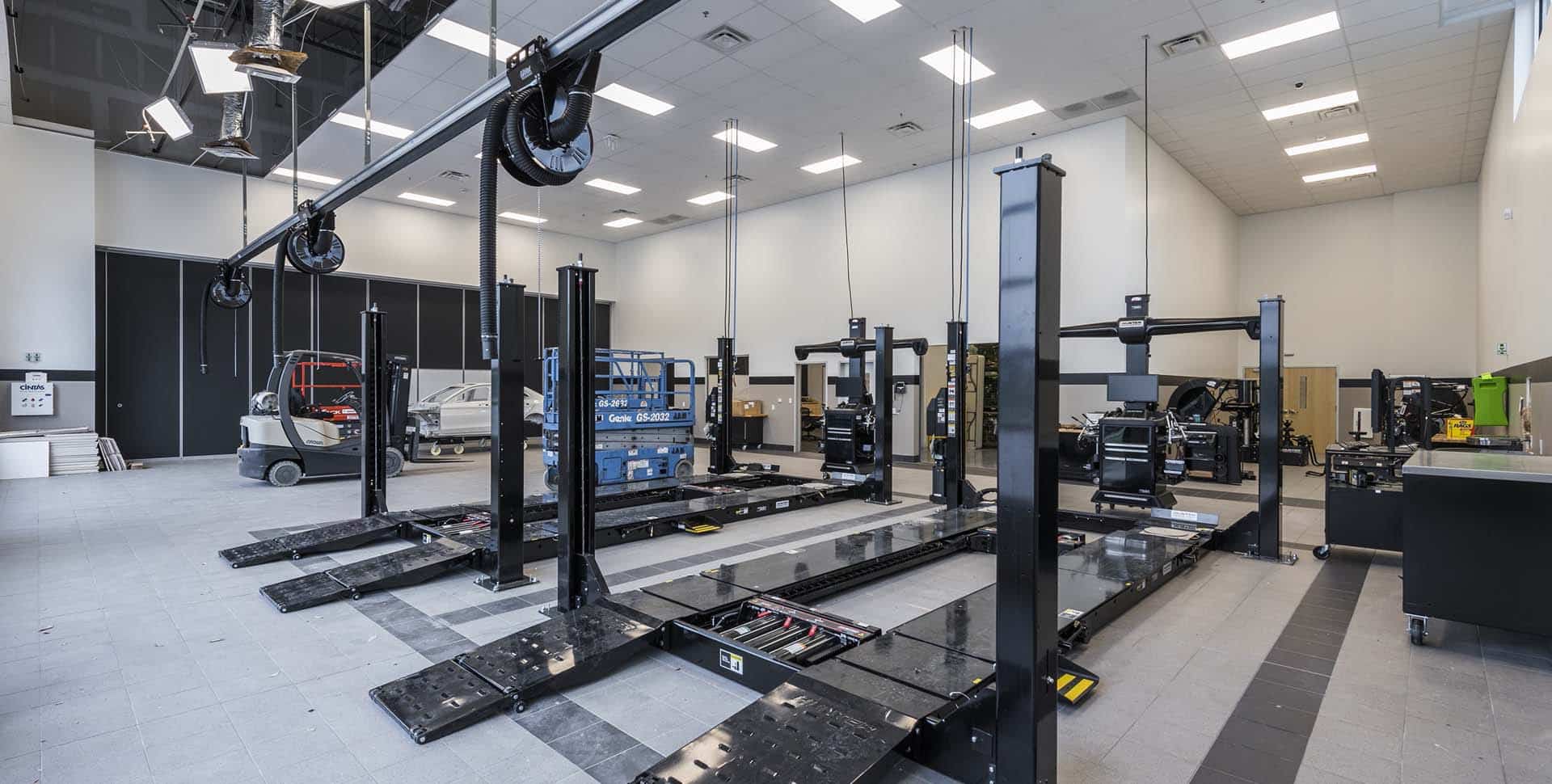 Large automotive workshop with multiple vehicle lifts, equipment, and workstations, illuminated by overhead lighting in a spacious, clean environment.