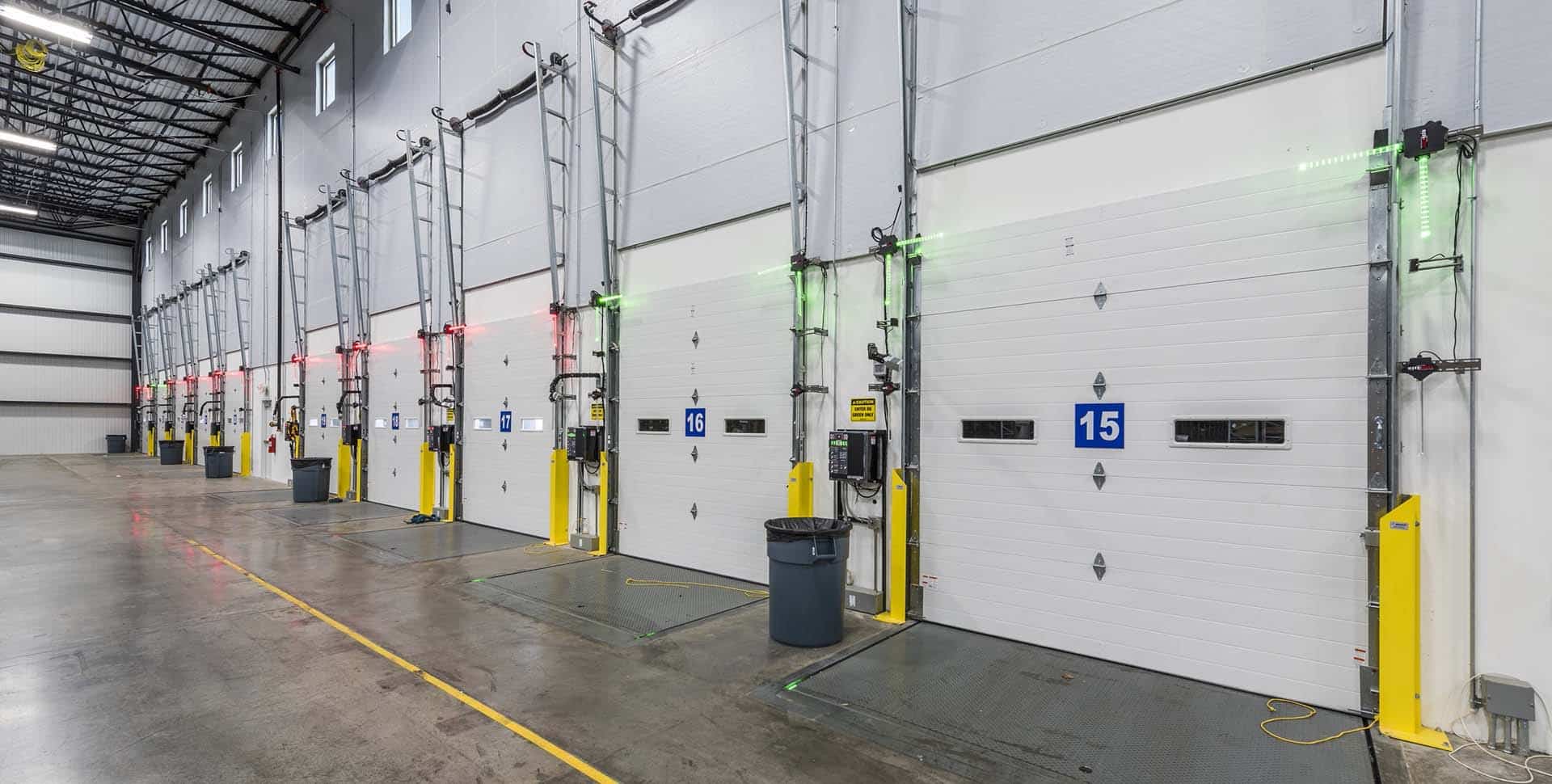 Interior view of a warehouse loading dock with multiple closed bay doors, each labeled with numbers and equipped with green and red indicator lights.