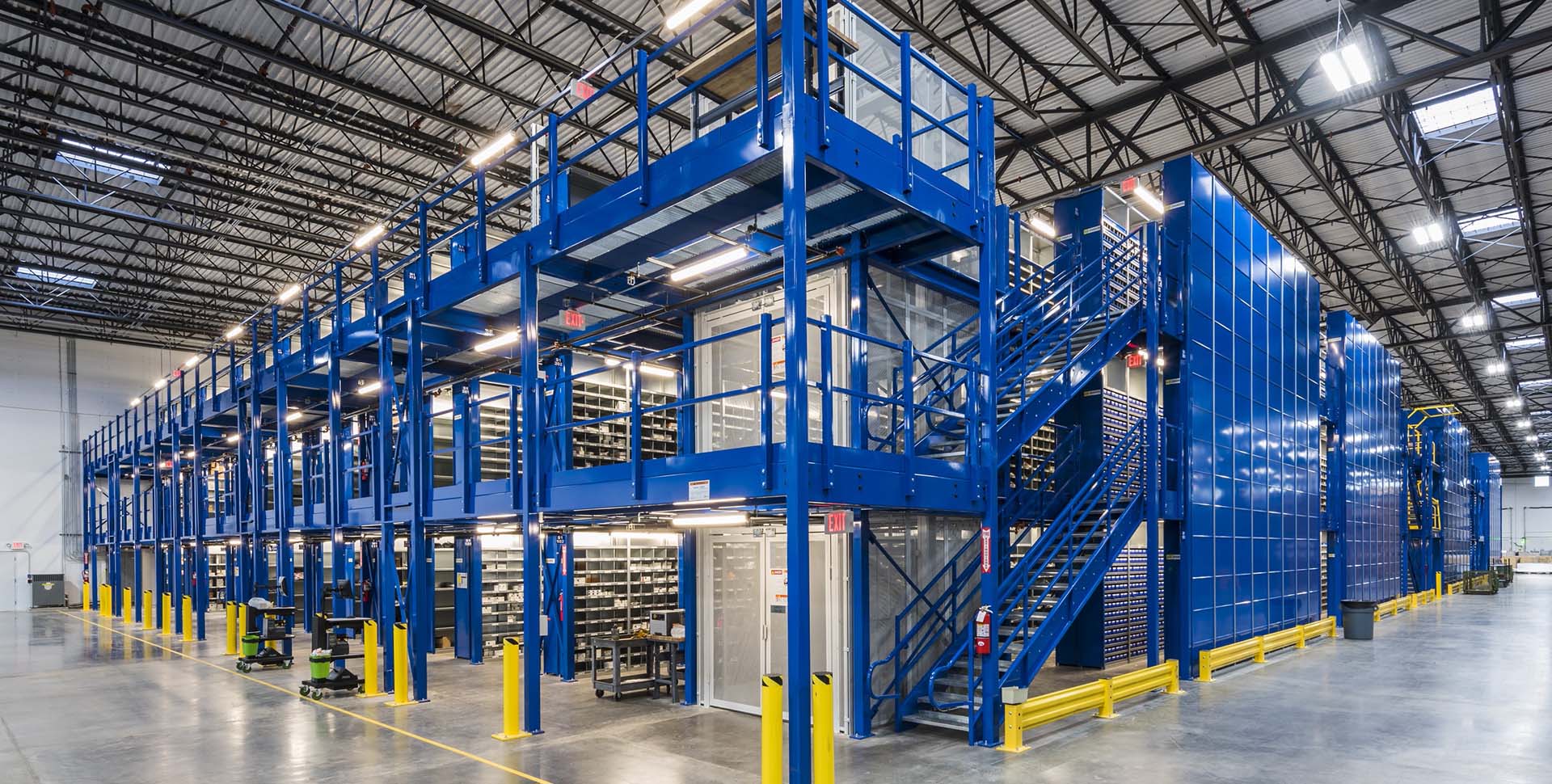 Large industrial warehouse interior with a blue metal mezzanine structure, stairs, shelving units, and bright overhead lighting.