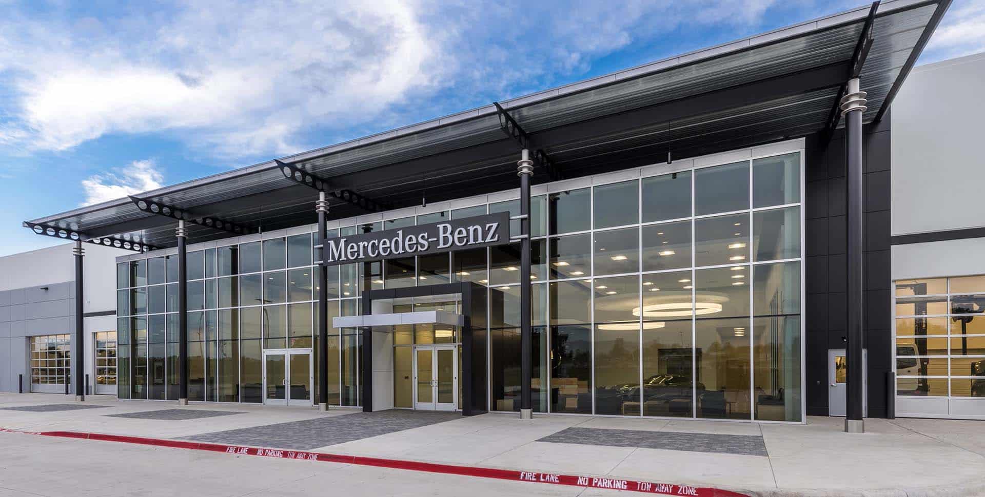 Modern glass-fronted Mercedes-Benz dealership building with large windows, metal awning, and the brand's sign above the main entrance under a partly cloudy sky.