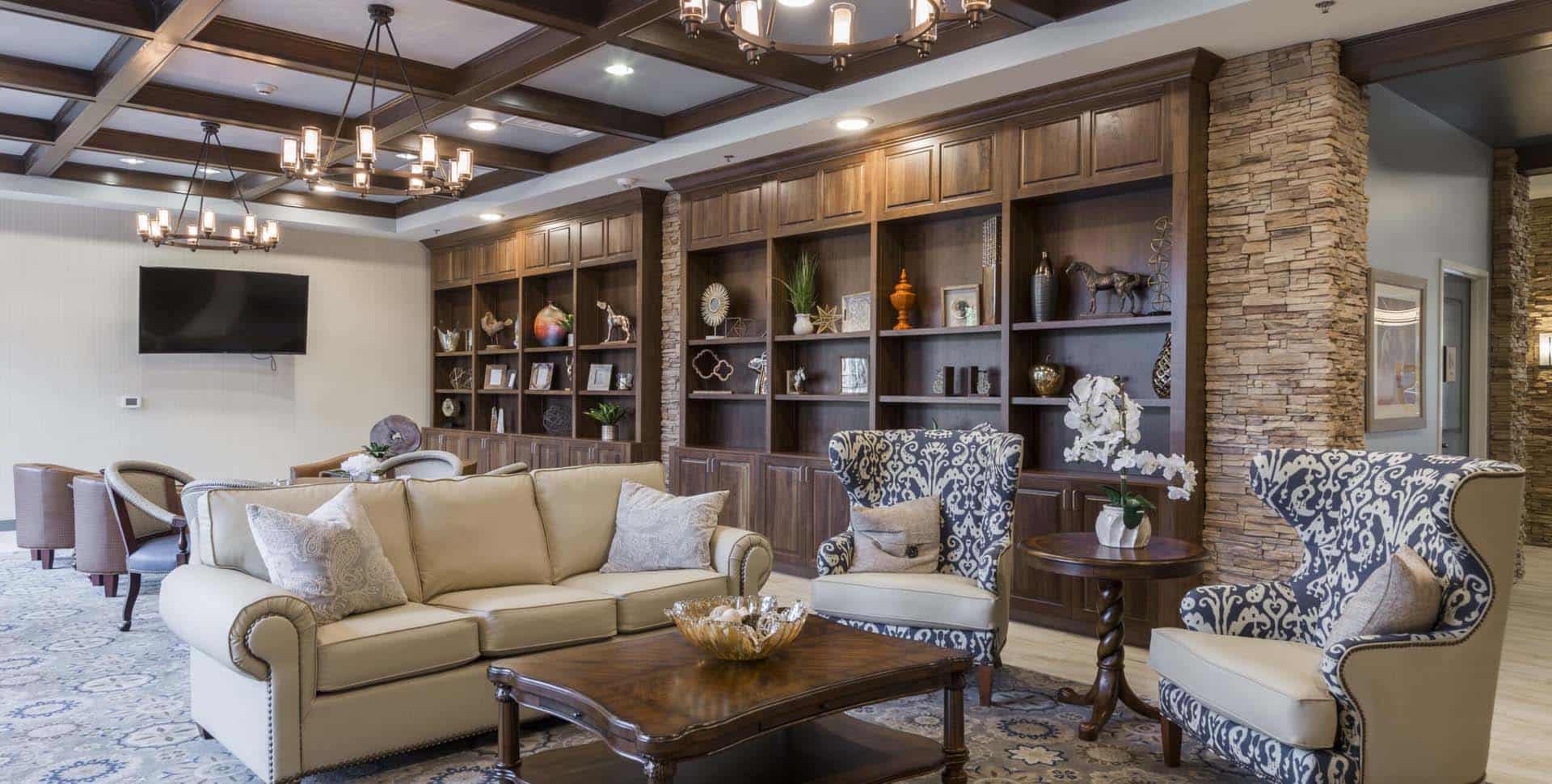 A spacious living room with cream sofas, patterned armchairs, a wooden coffee table, built-in bookshelves, and stone accent walls.