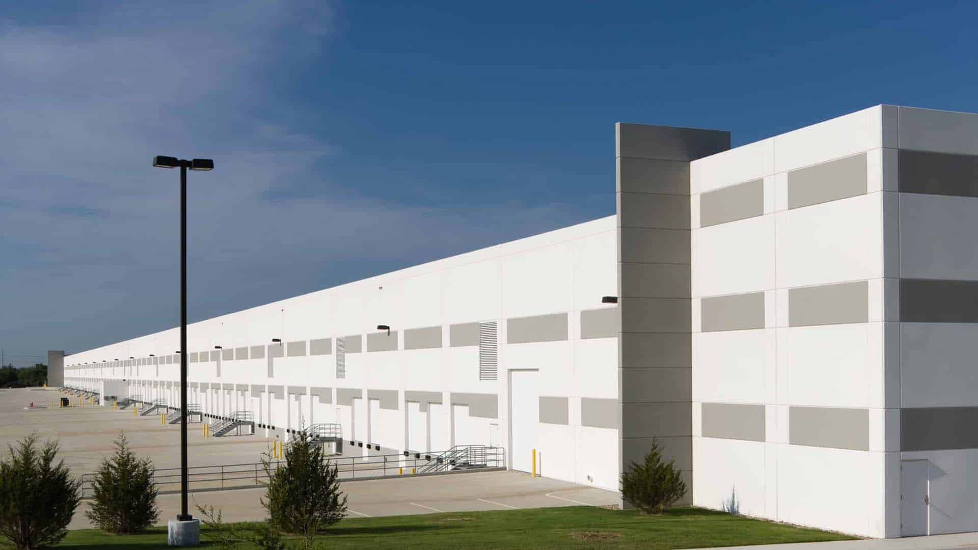 Large white industrial warehouse with multiple loading docks, a clear blue sky, and sparse landscaping in the foreground.