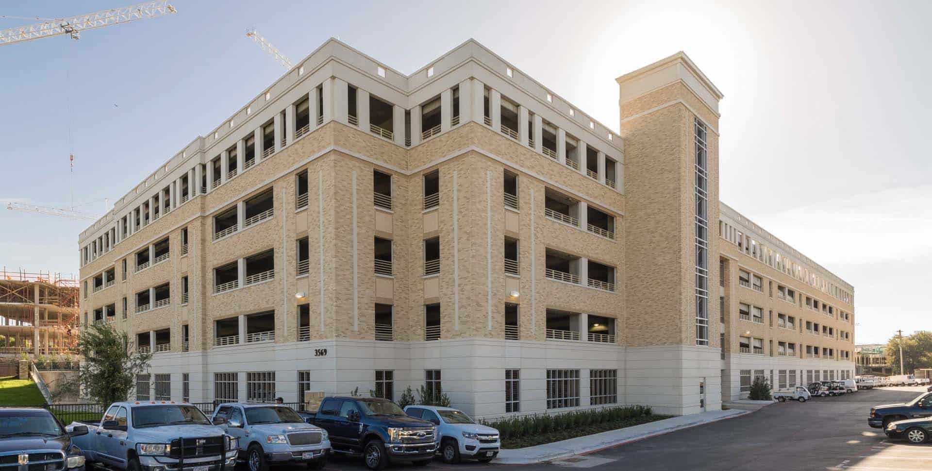 Large multi-level parking garage made of tan brick and concrete, with several parked vehicles in front and a clear sky overhead.