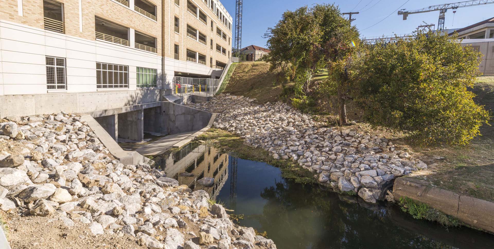 A small creek with rocky banks runs beside a modern building, with trees and grass on one side and construction cranes visible in the background.