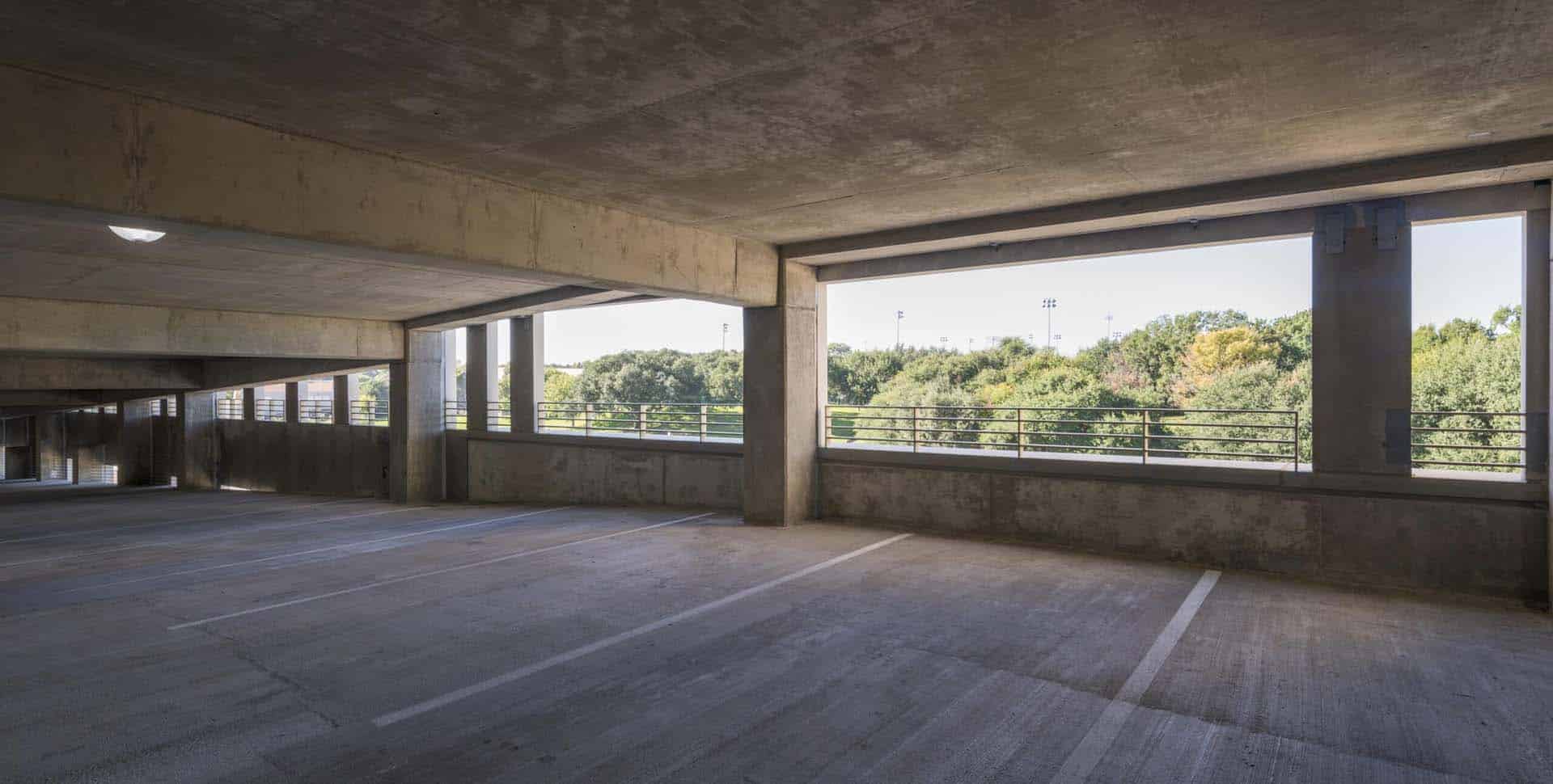 An empty concrete parking garage with sunlight coming through large openings, showing trees and greenery outside in the background.